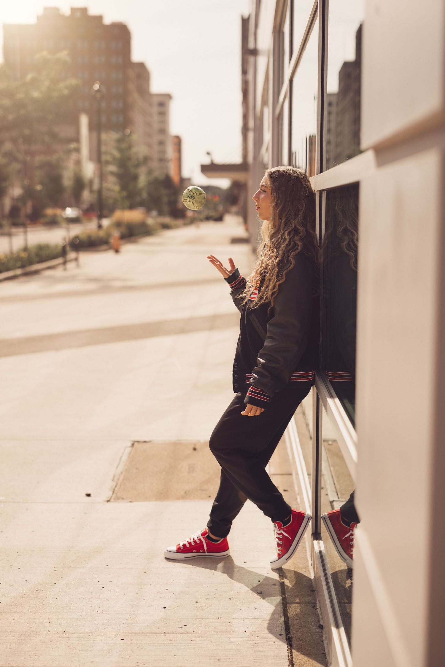 Person in black outfit and red sneakers poses playfully against reflective building windows.