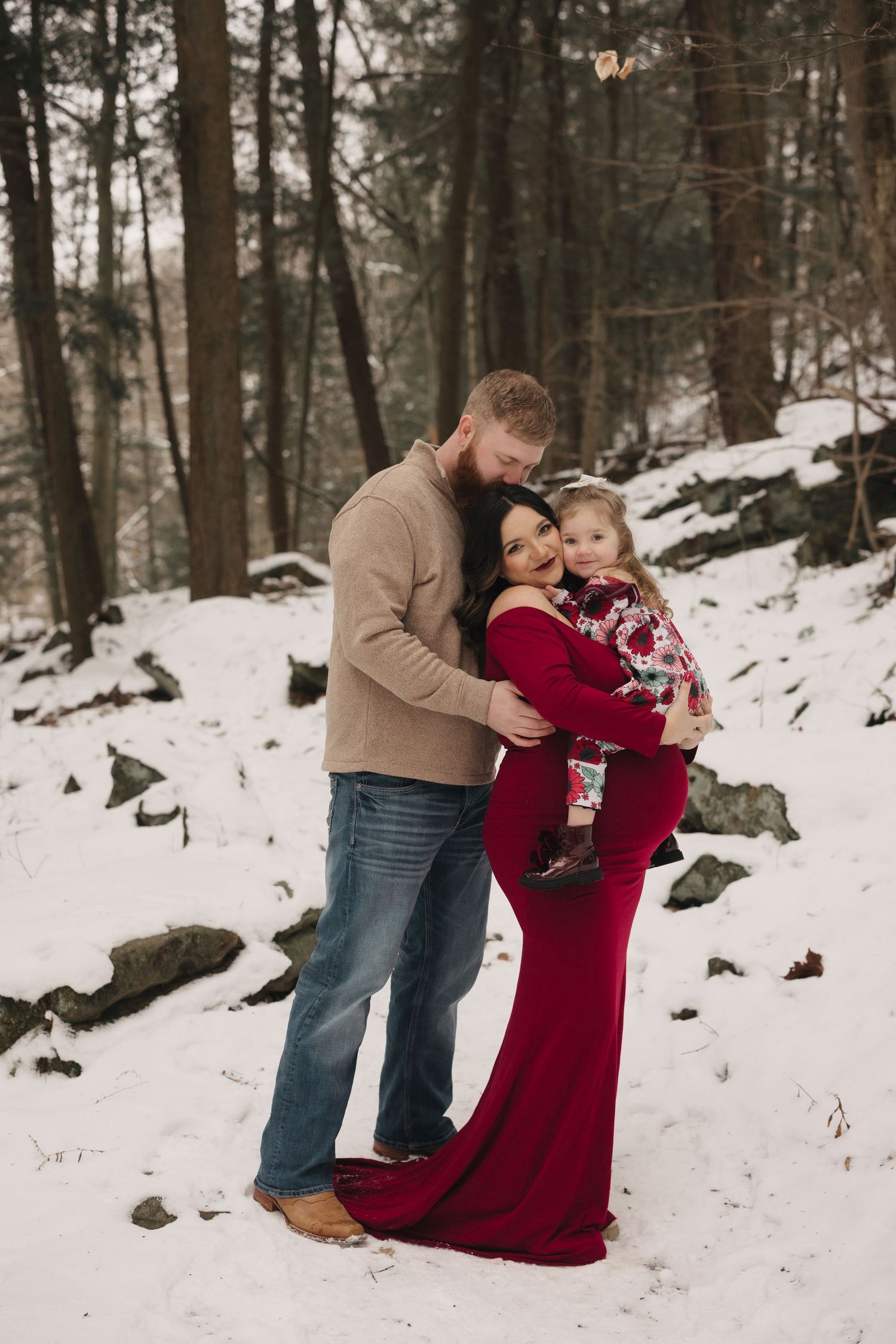 Winter family photoshoot in a snowy forest with burgundy dress against white landscape.