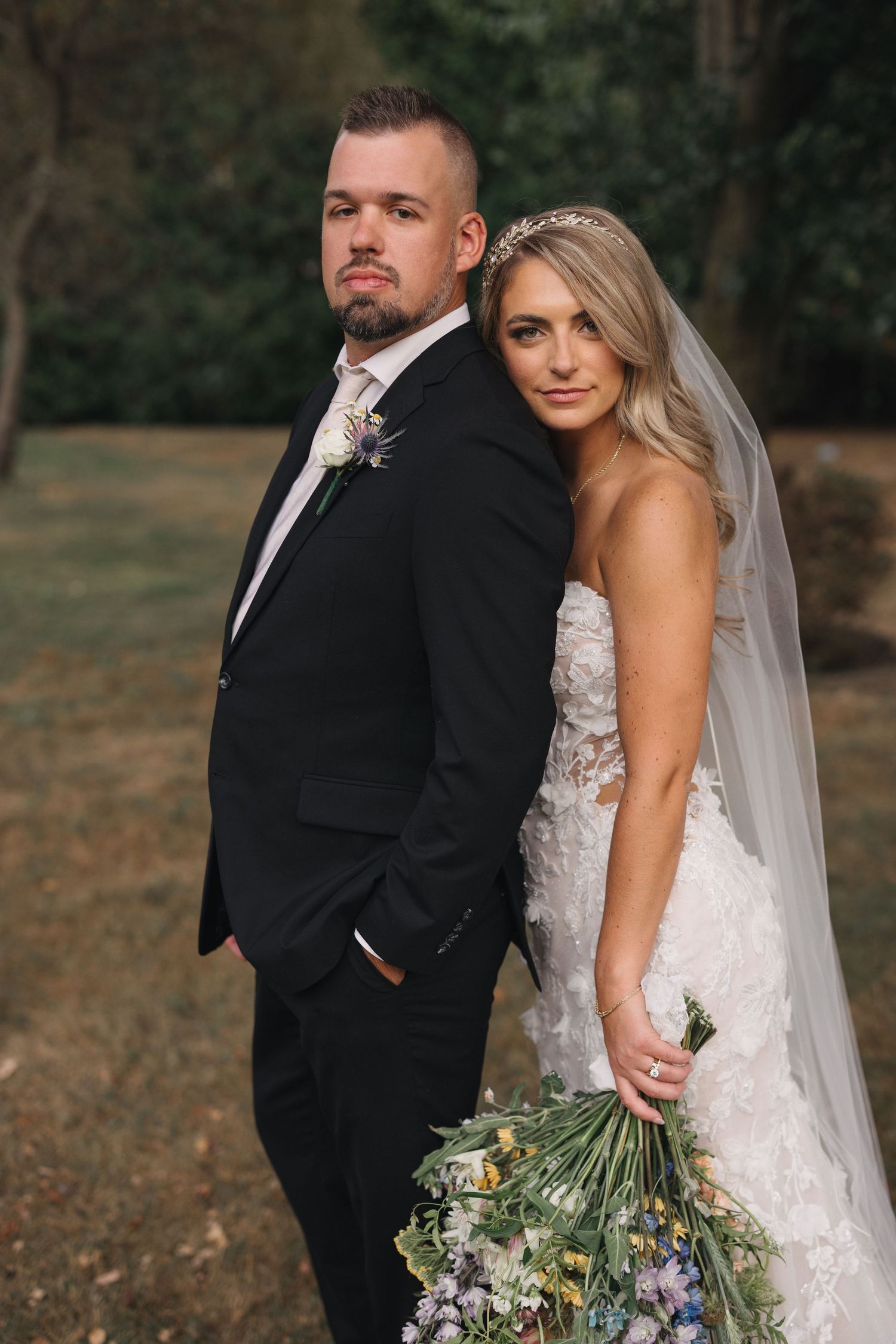 Wedding couple poses together outdoors with the bride holding a rustic floral bouquet.