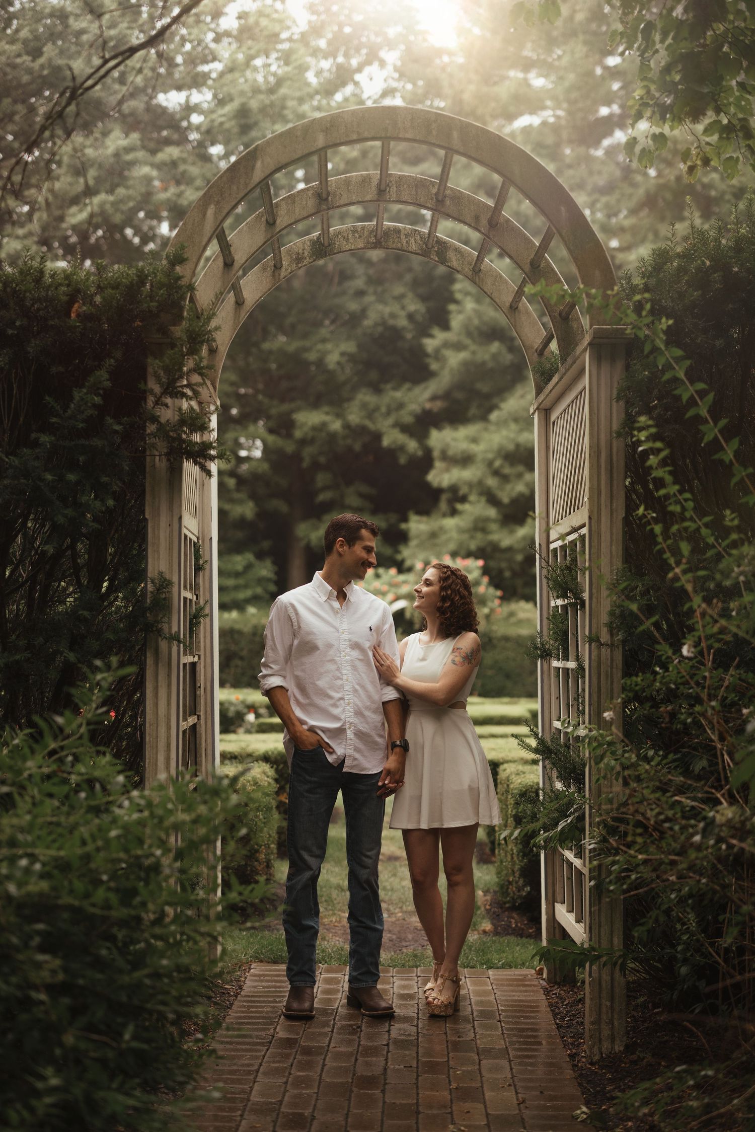 Romantic couple strolls through garden archway during golden hour photoshoot.