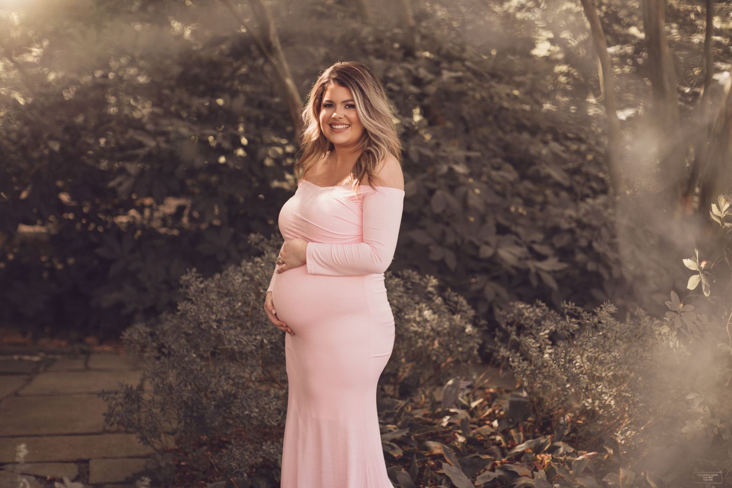 Maternity photo session in nature with soft pink dress and dreamy lighting.