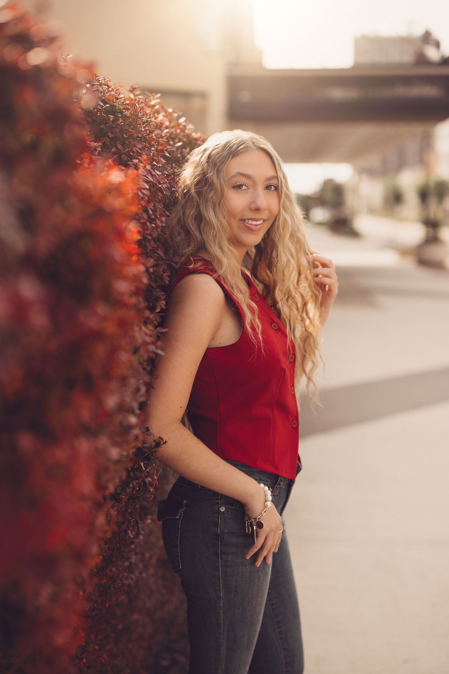 Woman in red sleeveless top poses against burgundy foliage background.