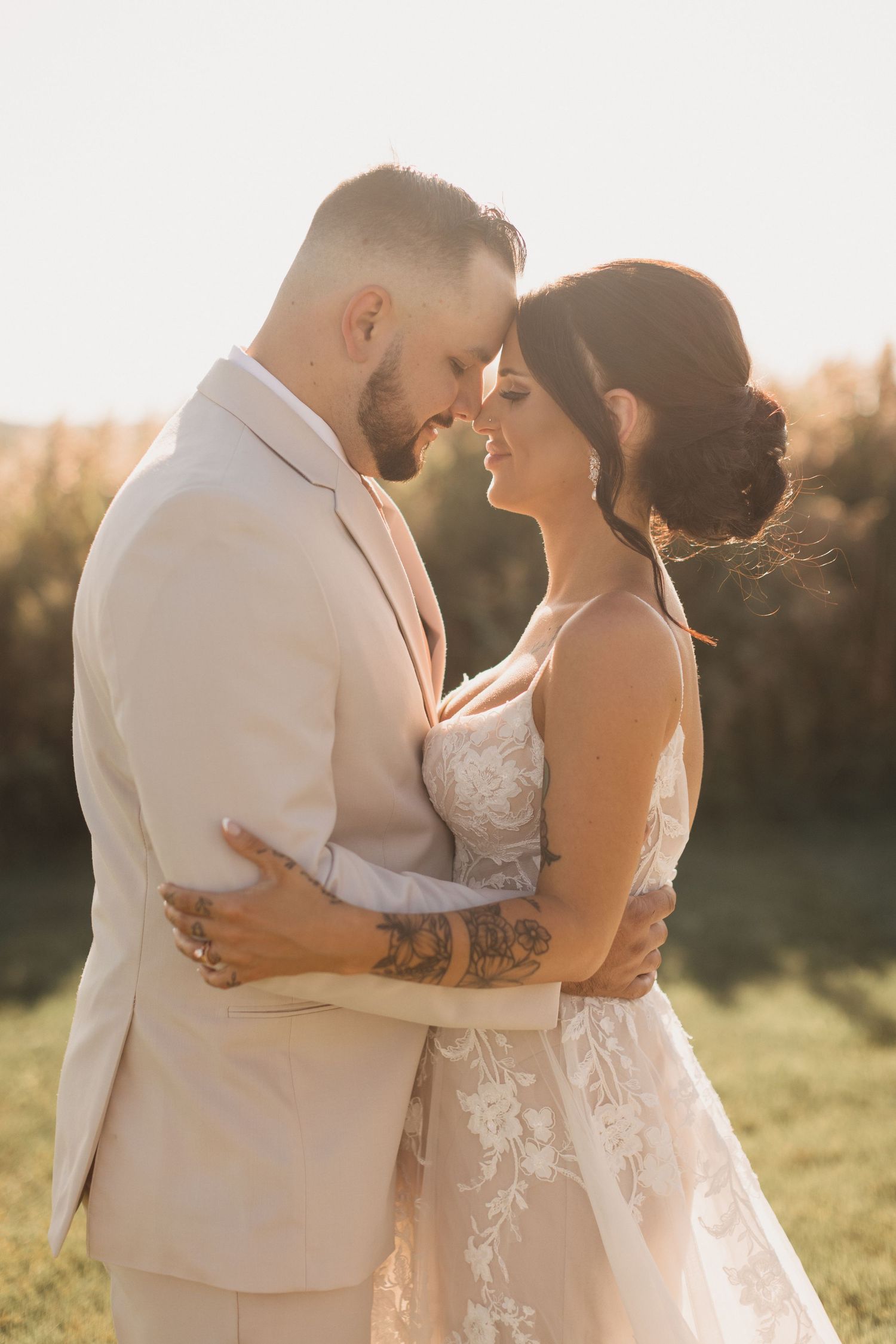 Wedding couple in light-colored attire embraces during golden hour in a grassy field.