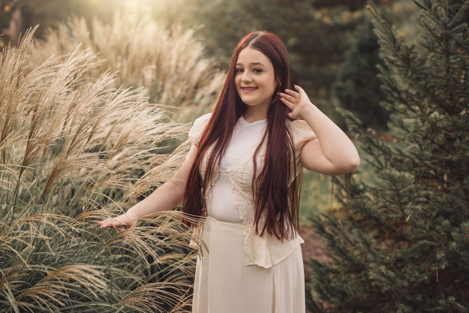 Woman in cream dress poses in natural outdoor setting with tall grass and sunlit backdrop.