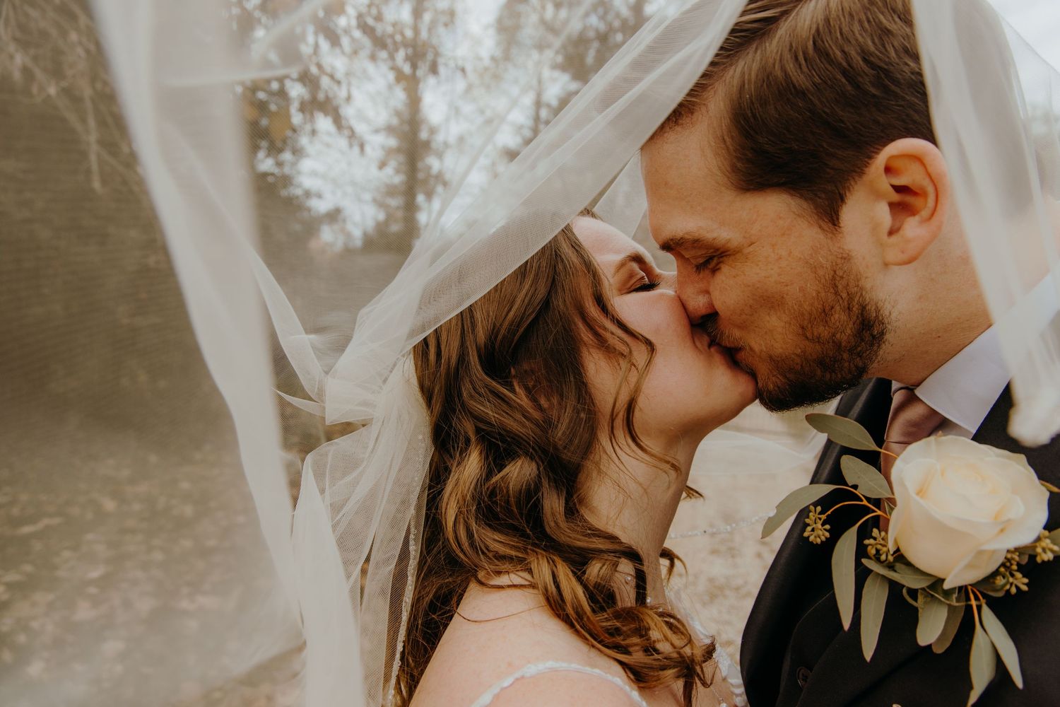 A romantic wedding couple shares a tender kiss under a flowing veil in soft, dreamy lighting.