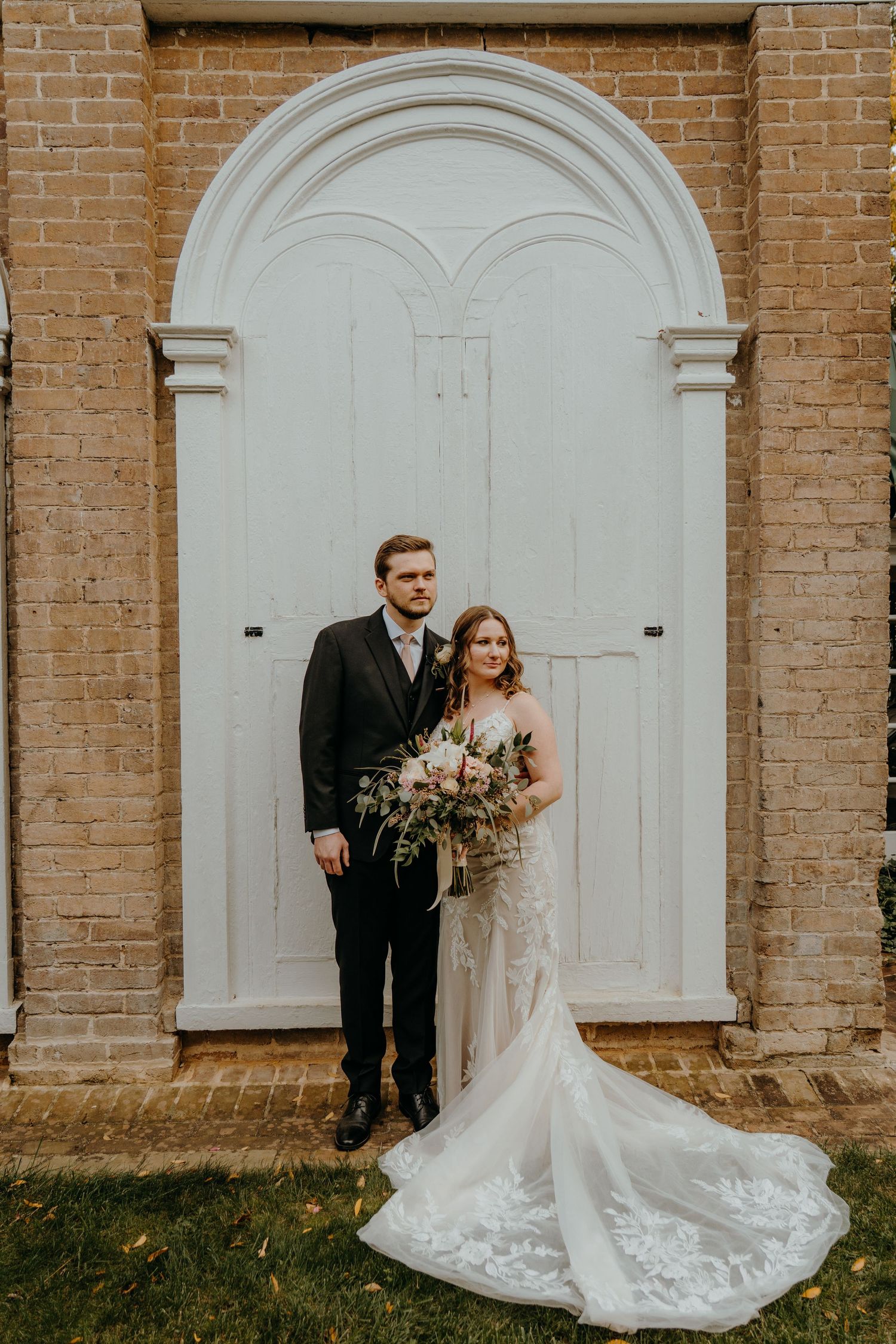 Elegant wedding couple poses against brick wall with white arched doorways in romantic outdoor setting. Wedding at the Preserve at Dundee