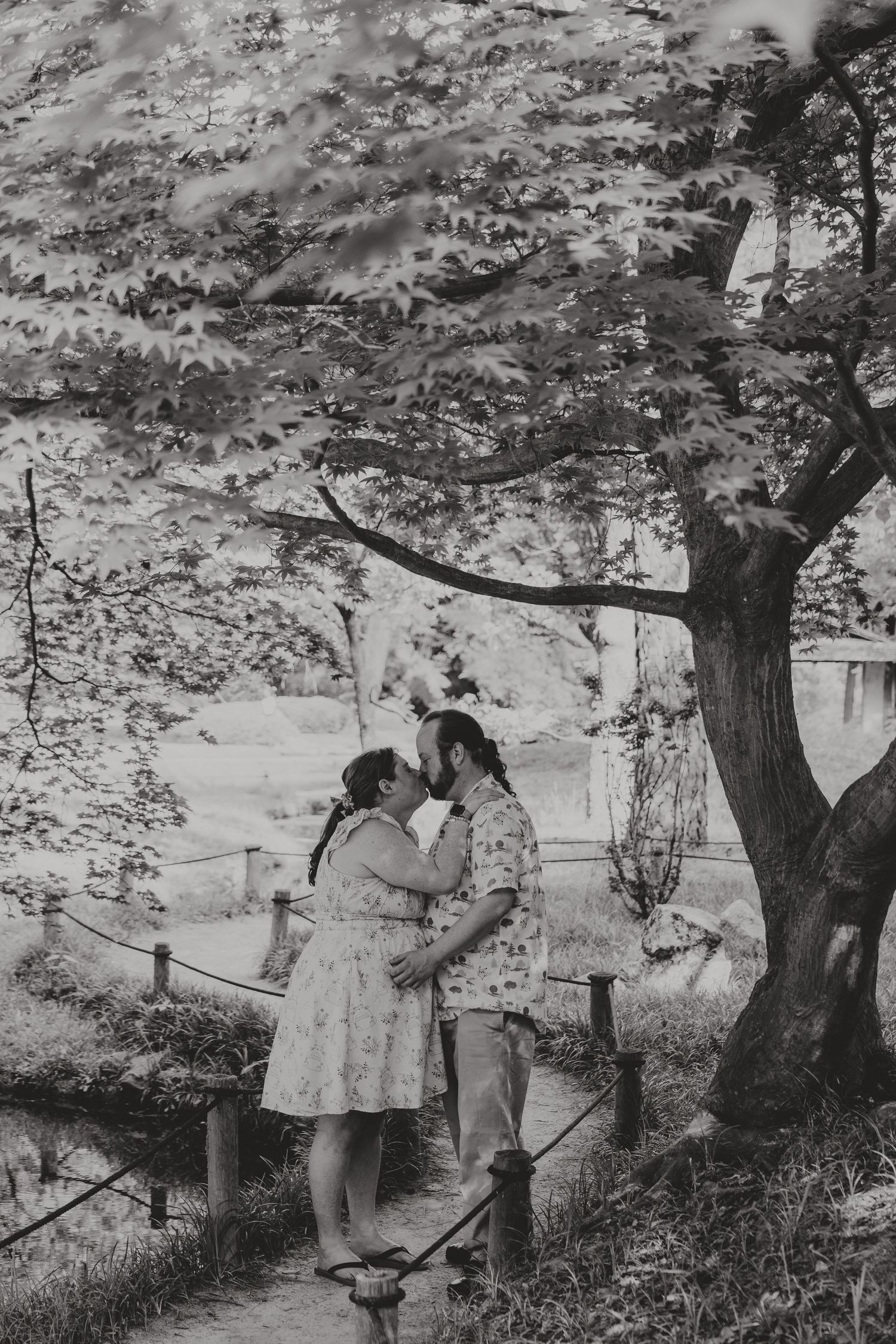 Black and white photo of a couple sharing a romantic moment under a large tree in a lush garden setting.