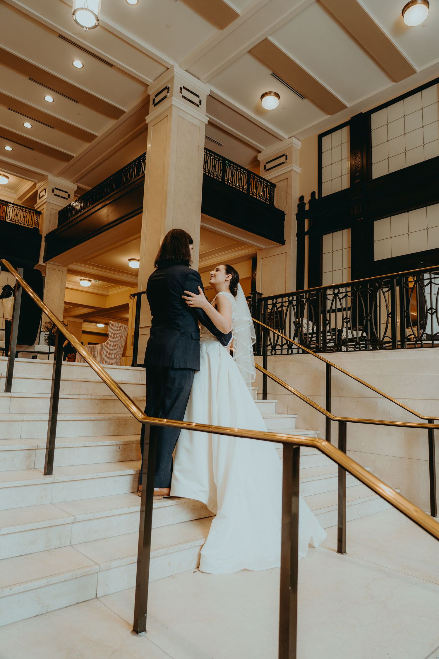 Bride and groom embrace on elegant marble staircase with gold railings in a grand historic wedding venue.