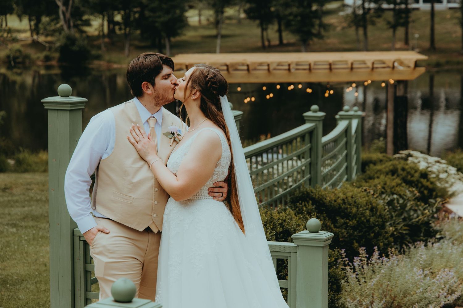 A bride and groom share a romantic kiss outdoors near a scenic pond with a pergola and string lights in the background.