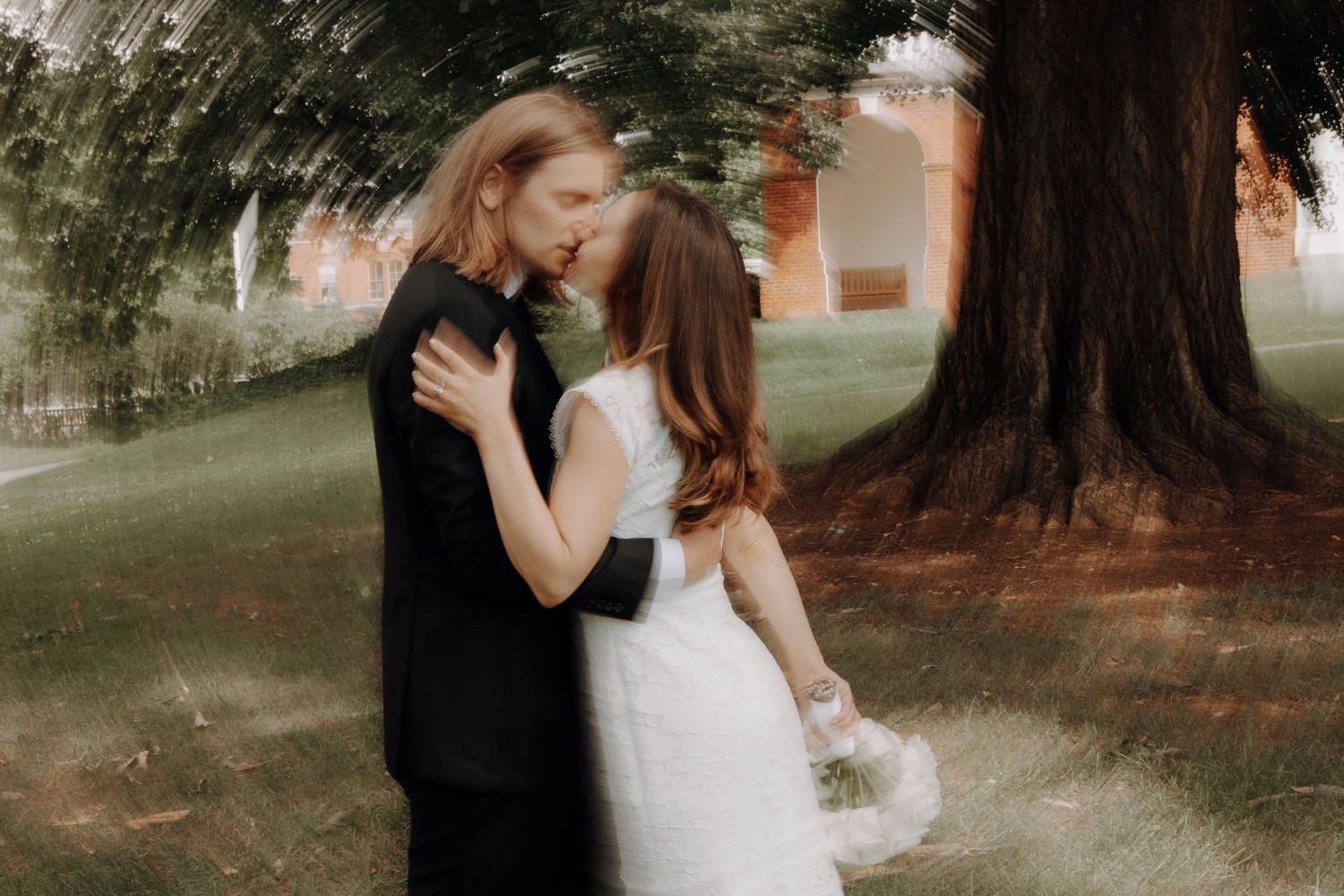 A romantic wedding couple embraces under a large tree in a dreamy outdoor setting.