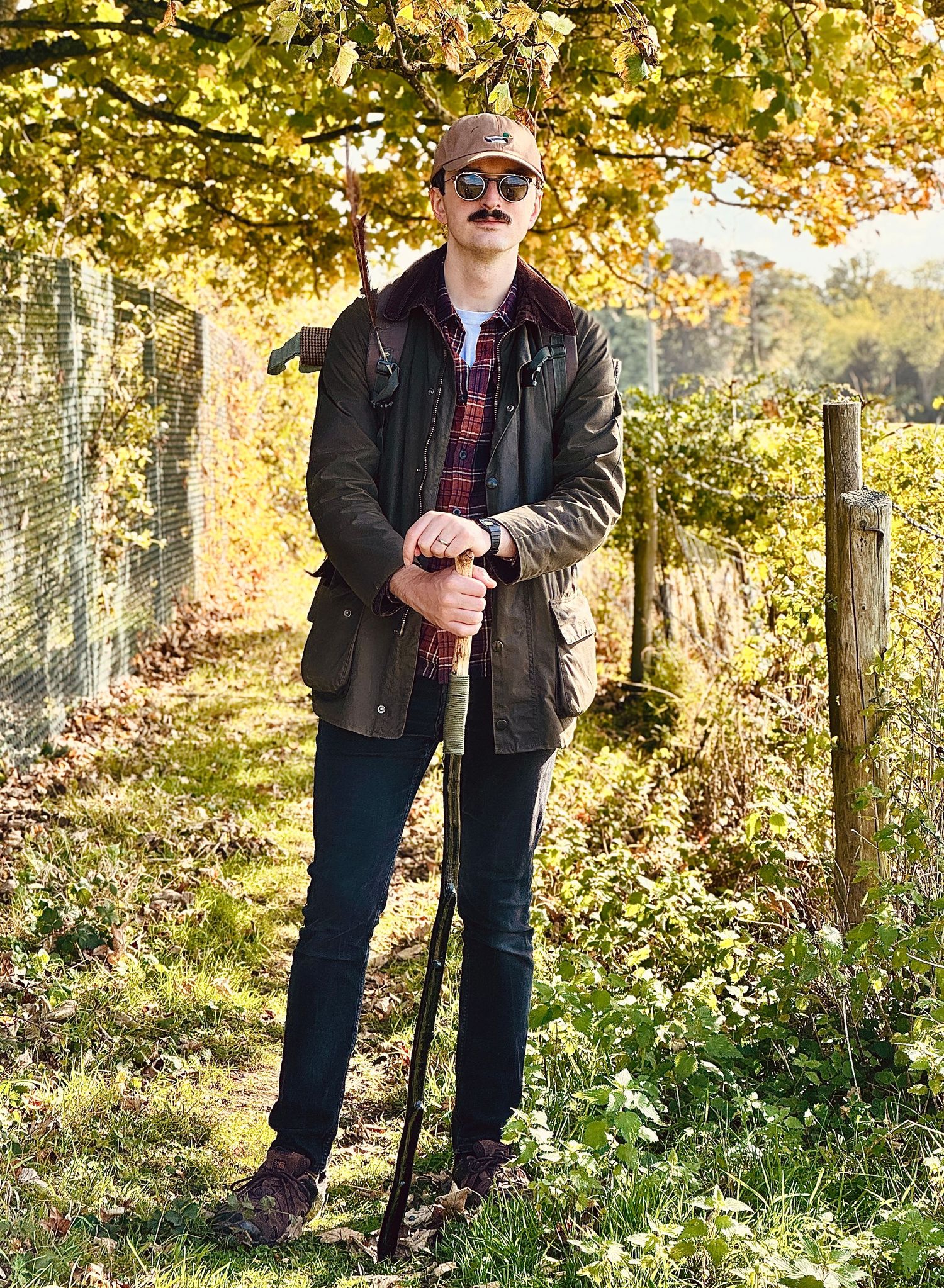 Someone stands with a walking stick in an autumn garden surrounded by golden leaves and tall trees.