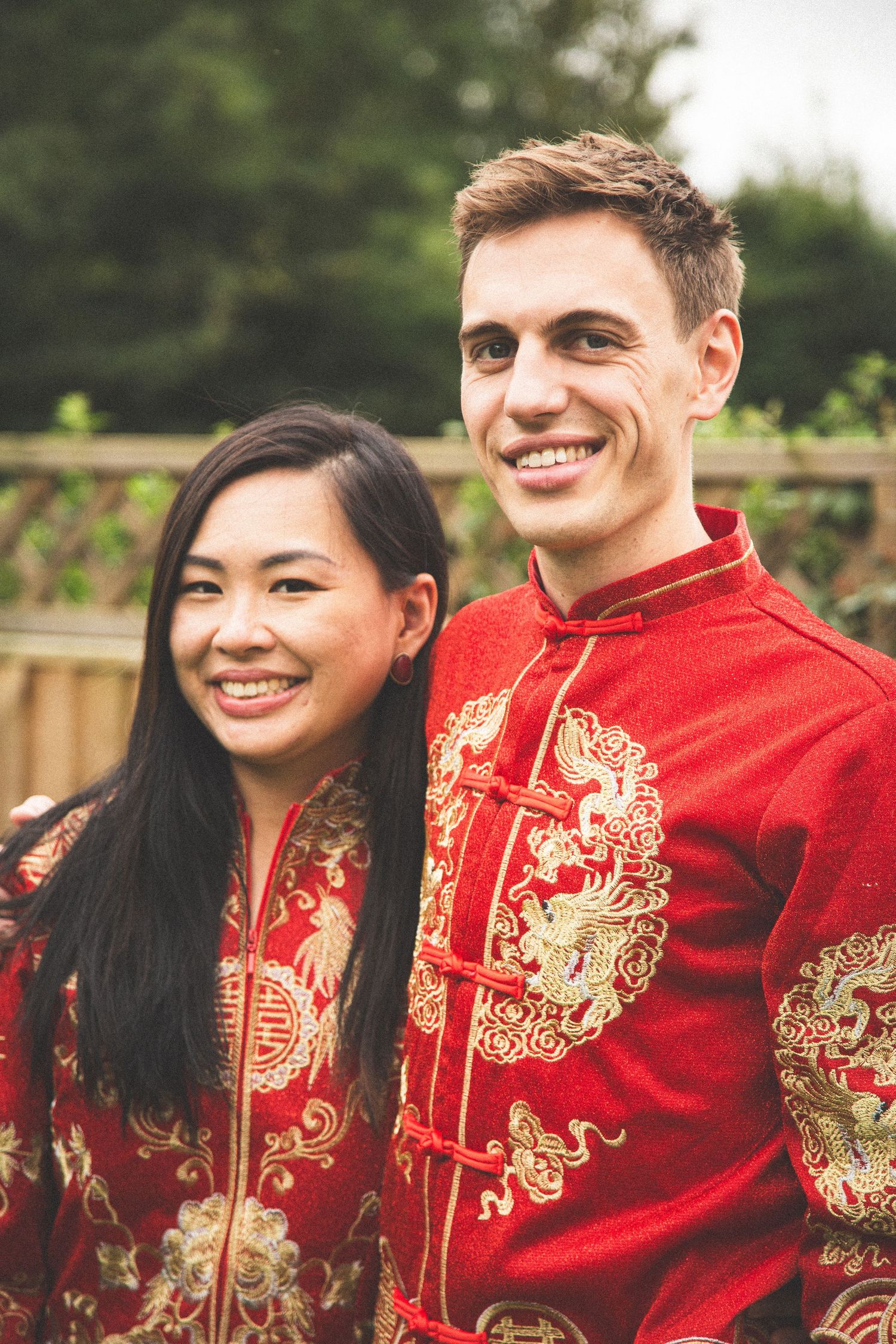 Couple wearing traditional red and gold Chinese wedding attire smile together in outdoor garden setting.