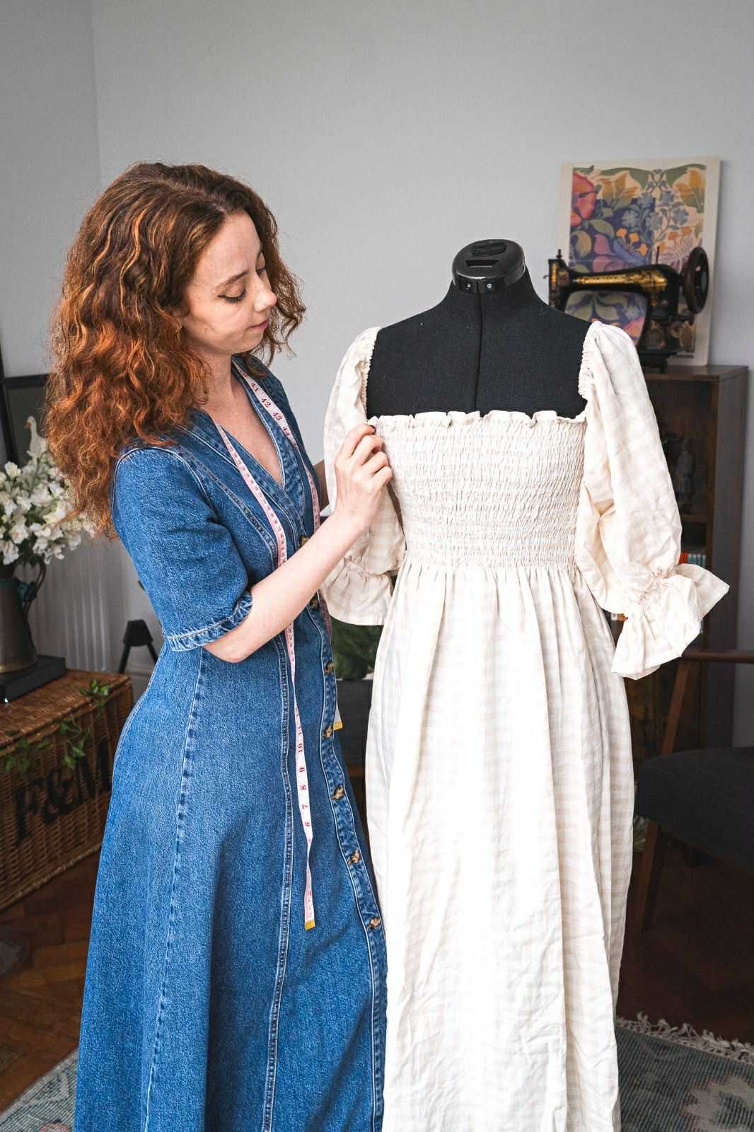 A woman in a denim dress adjusts a white Victorian-style dress on a mannequin form in a home setting.