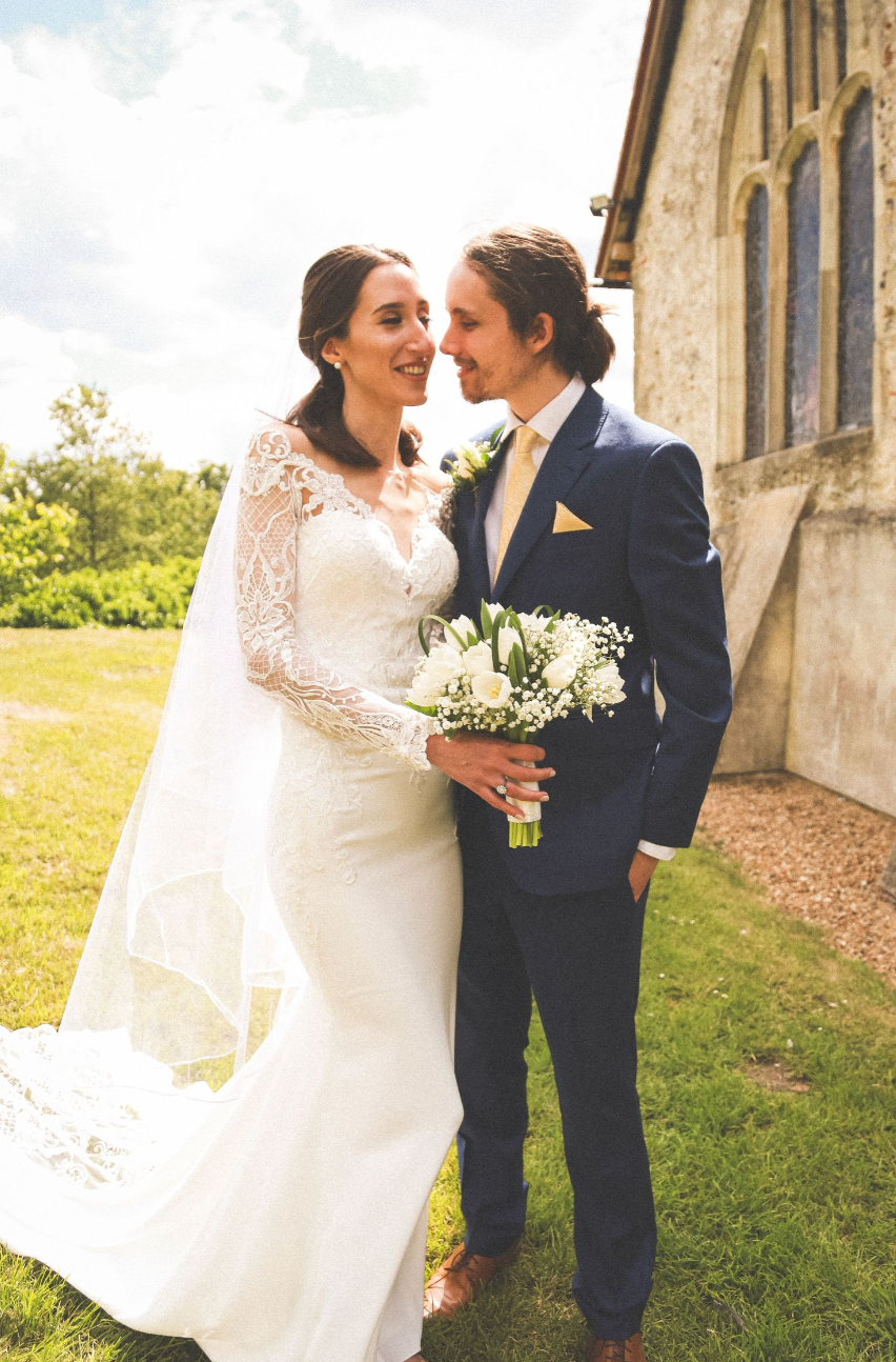 A happy couple shares a joyful moment together outside a church on their wedding day, holding a white floral bouquet.