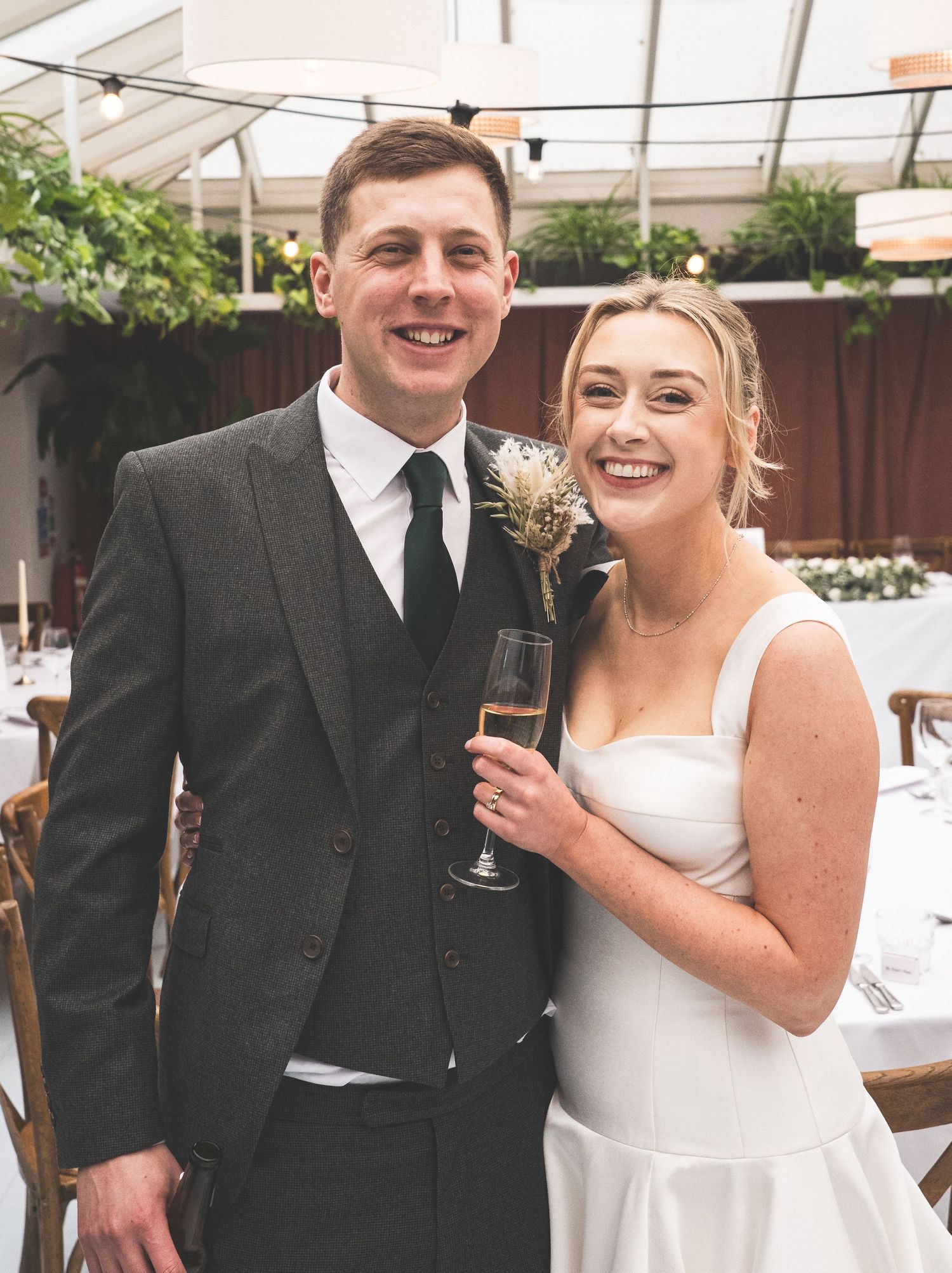 A smiling bride and groom pose together at their wedding reception, she holds a champagne flute wearing a white dress.