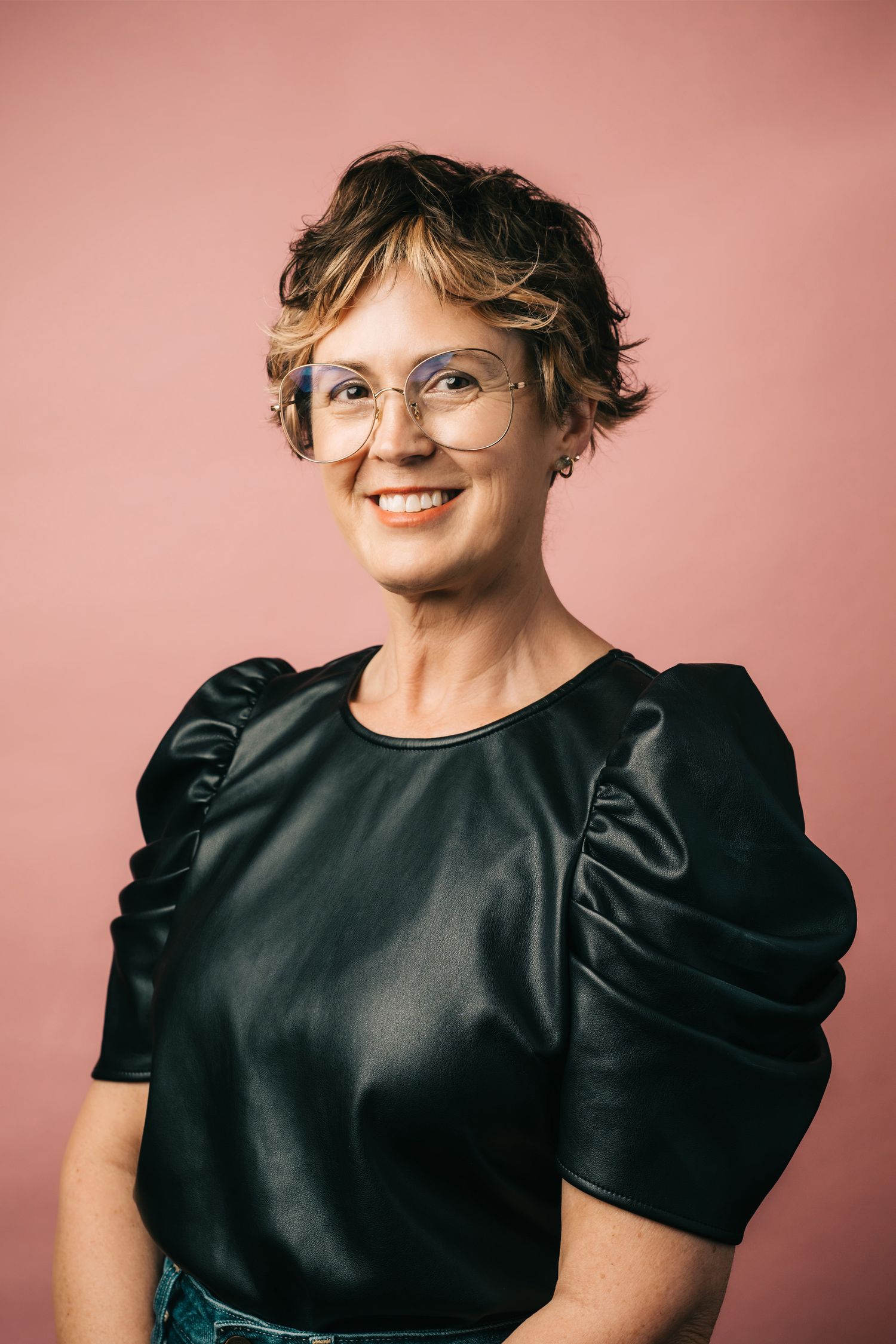 Portrait of someone in a black satin puff-sleeve top and glasses against a pink background.