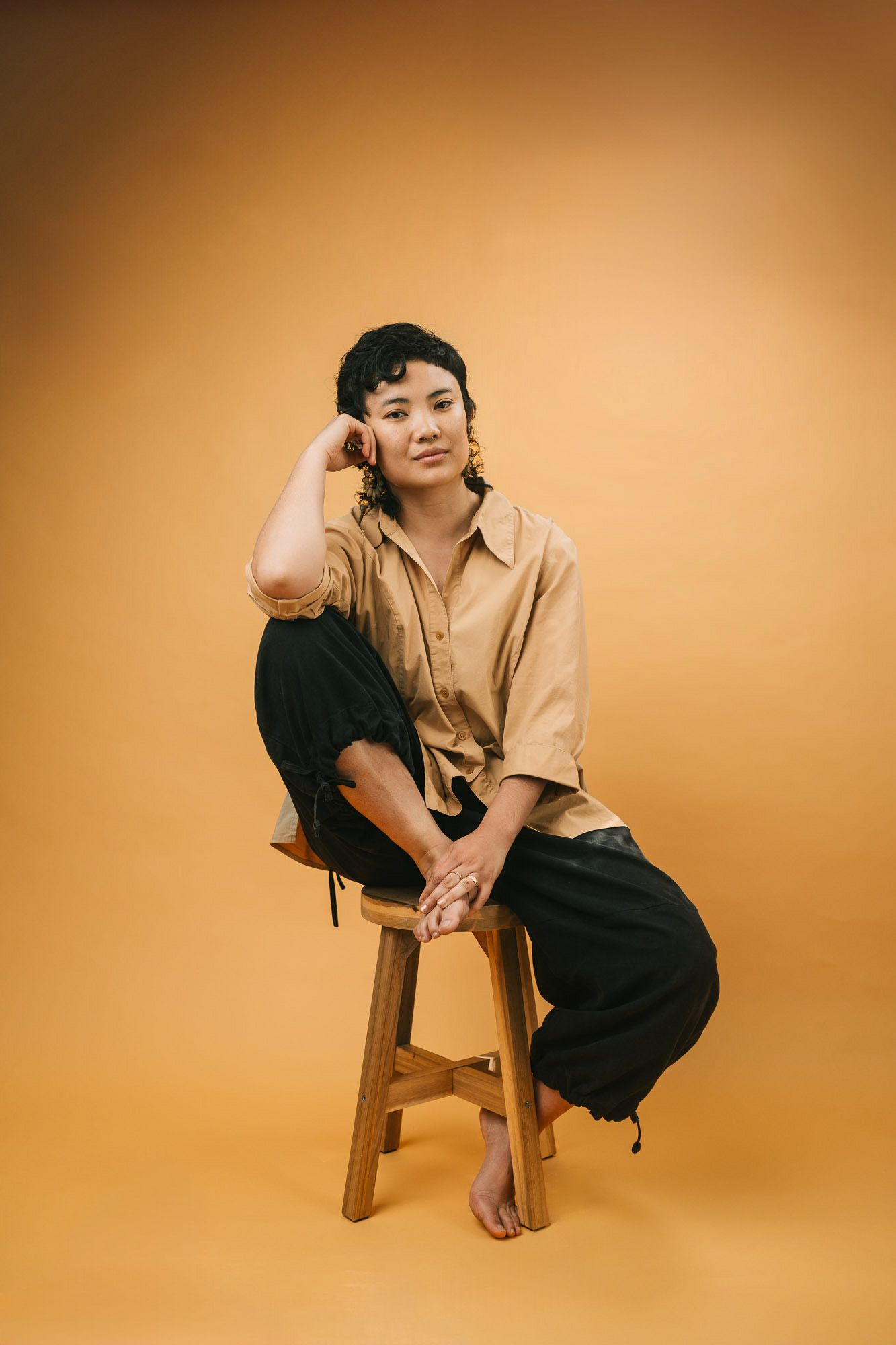 Casual portrait on wooden stool against warm orange background in relaxed styling.