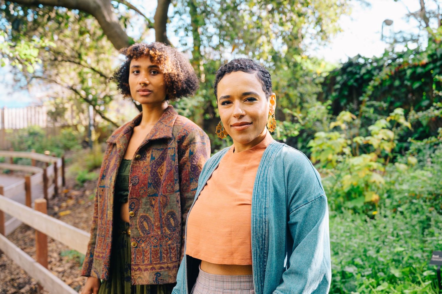 Candid shot of two friends in patterned and denim outfits standing on wooden steps outdoors.