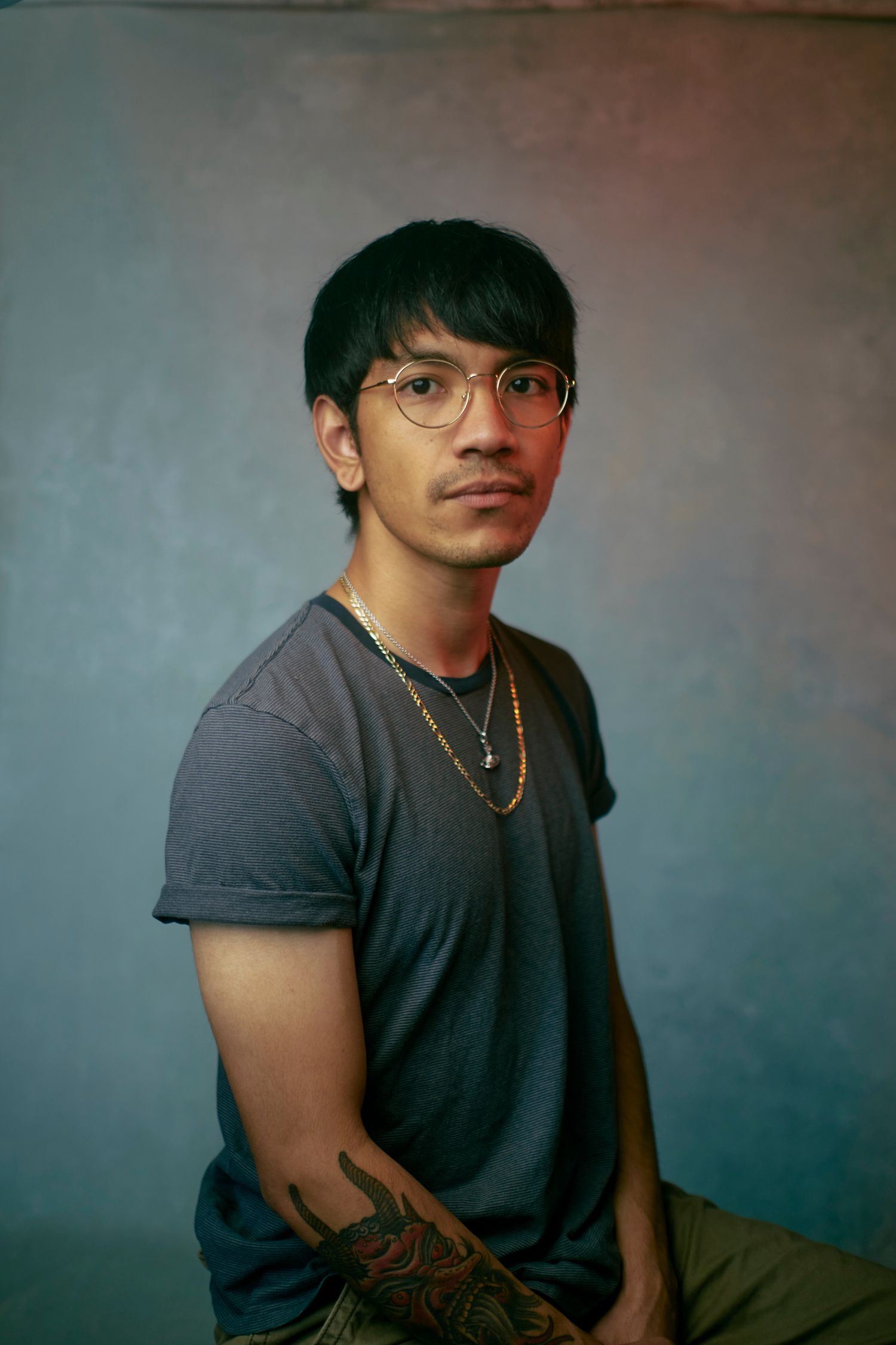 Portrait of a person in a gray t-shirt and glasses with tattoos visible against a gradient background.