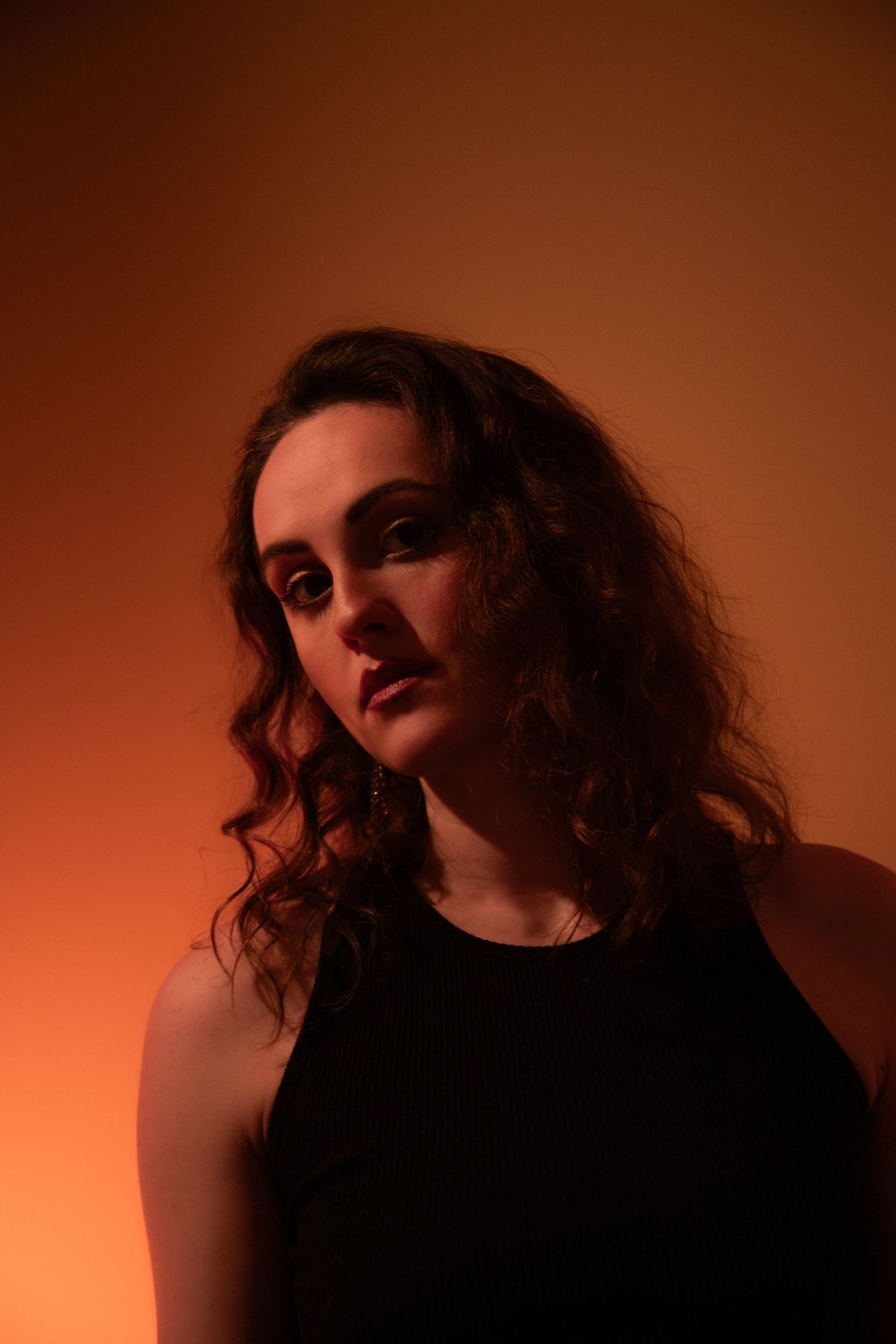 Woman with curly hair in black top poses against warm orange-red background in dramatic studio portrait.