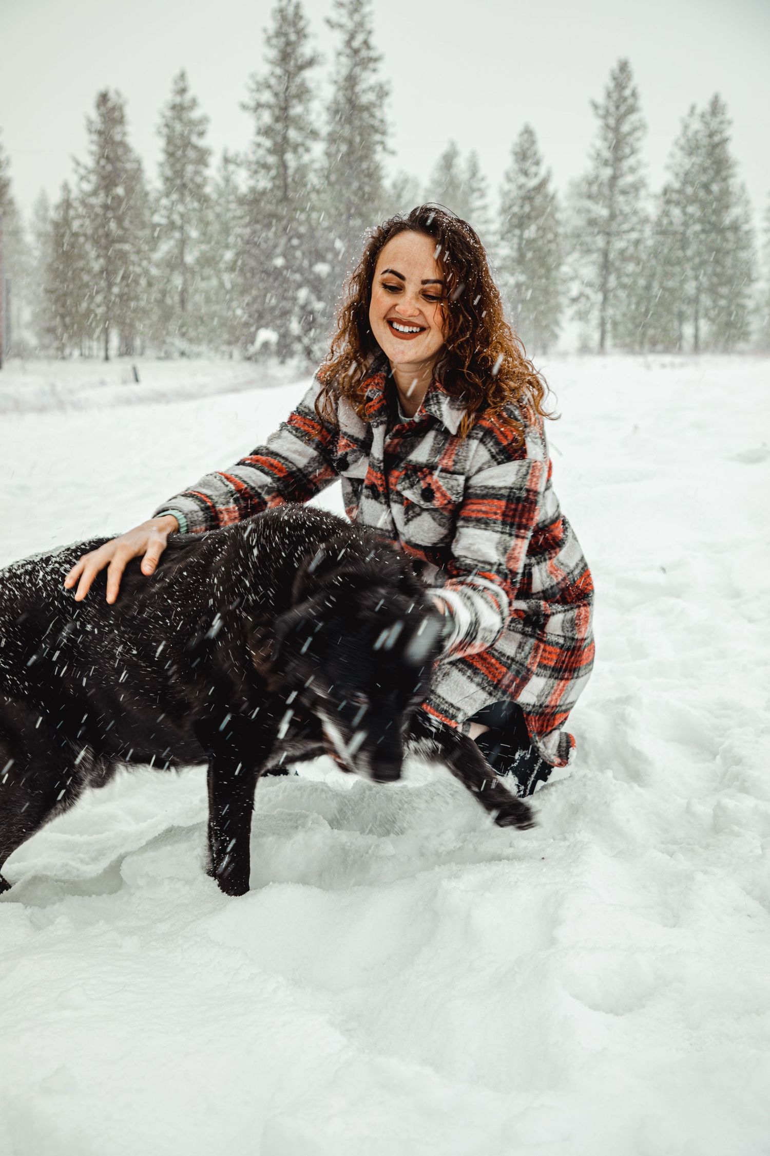 Person smiling while petting black dog in snowy forest setting.