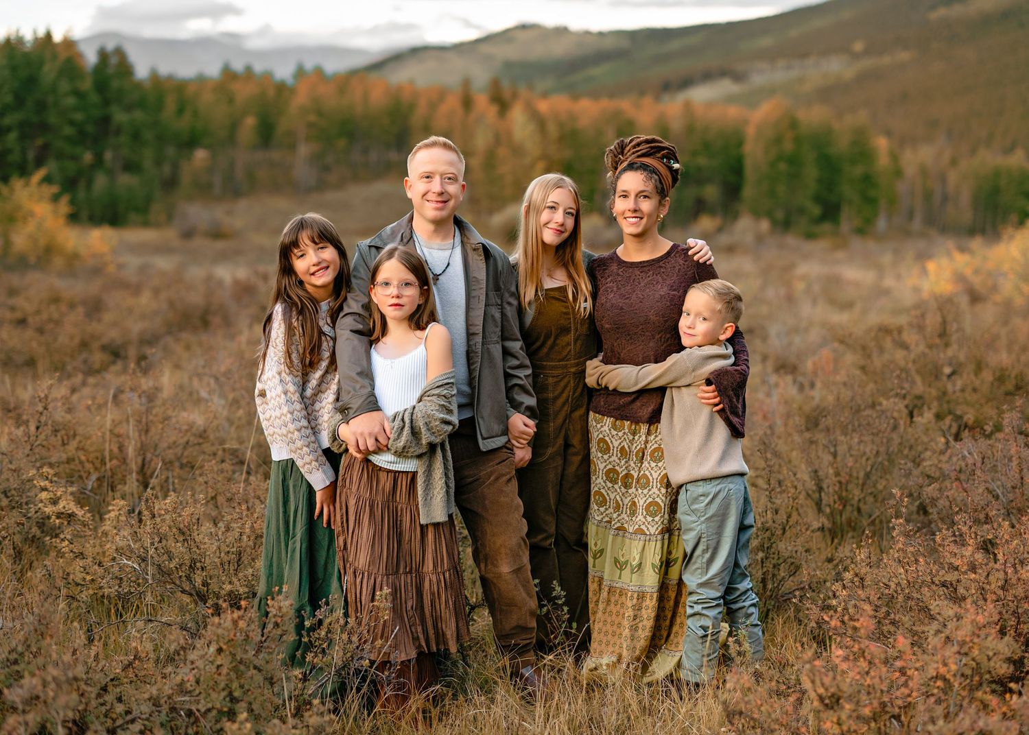 A group of people wearing earth-toned clothing stand together in a golden autumn field with mountains in the background.