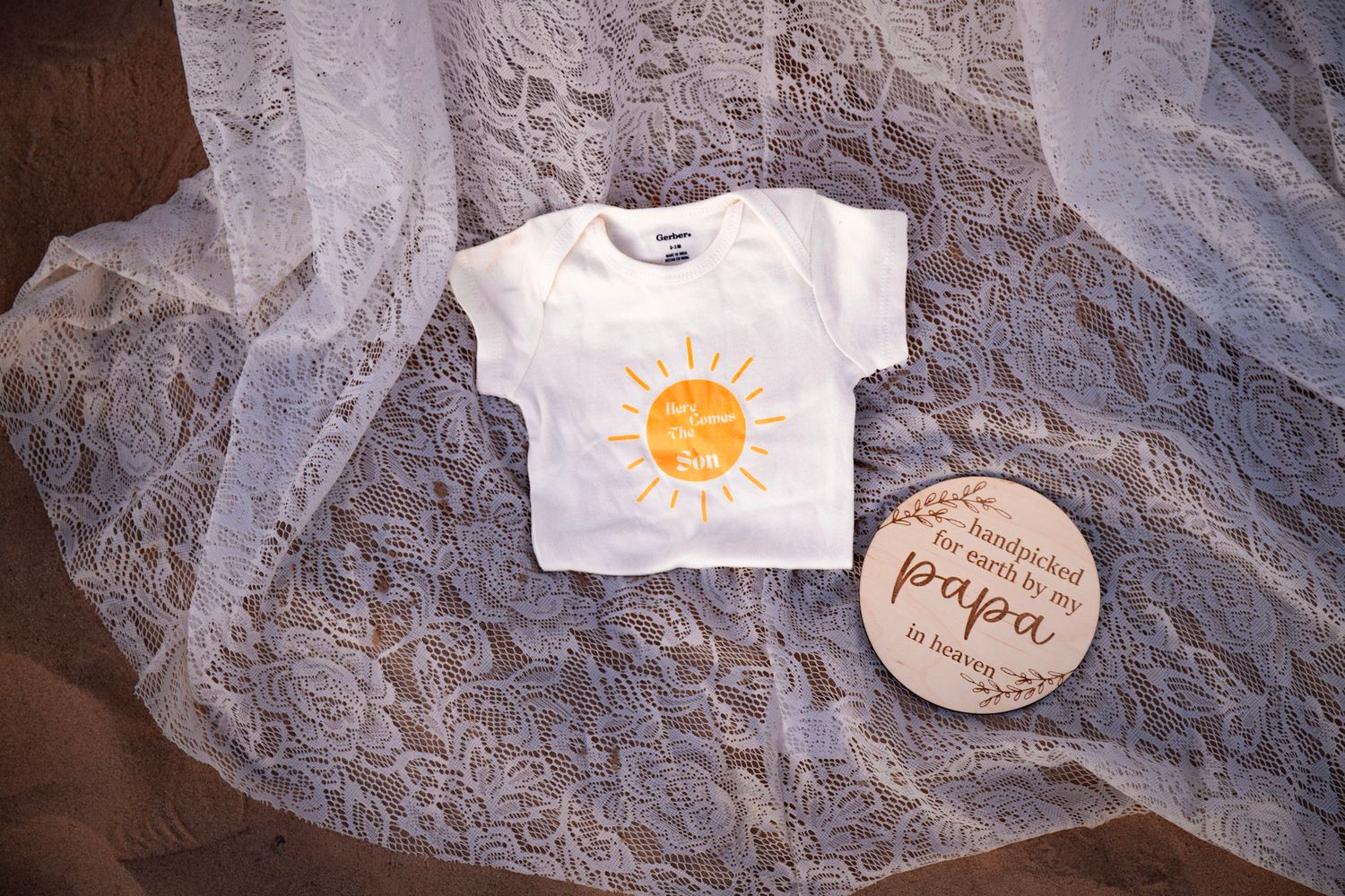 White baby onesie with sun print beside a wooden Papa sign on lace fabric, perfect for Father's Day gifting.