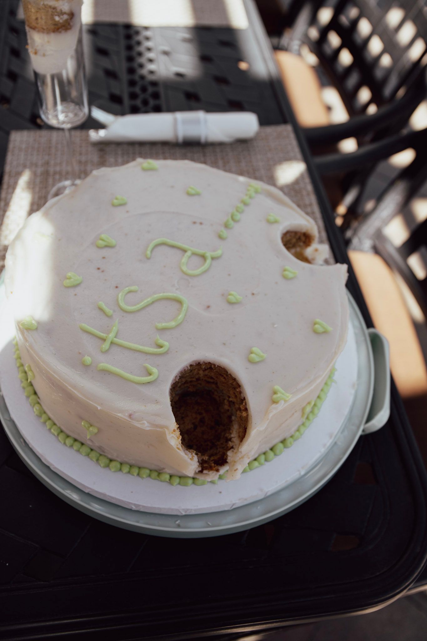 A partially eaten white cake with green frosting decorations reading 'it's 2' on a cake stand outdoors.