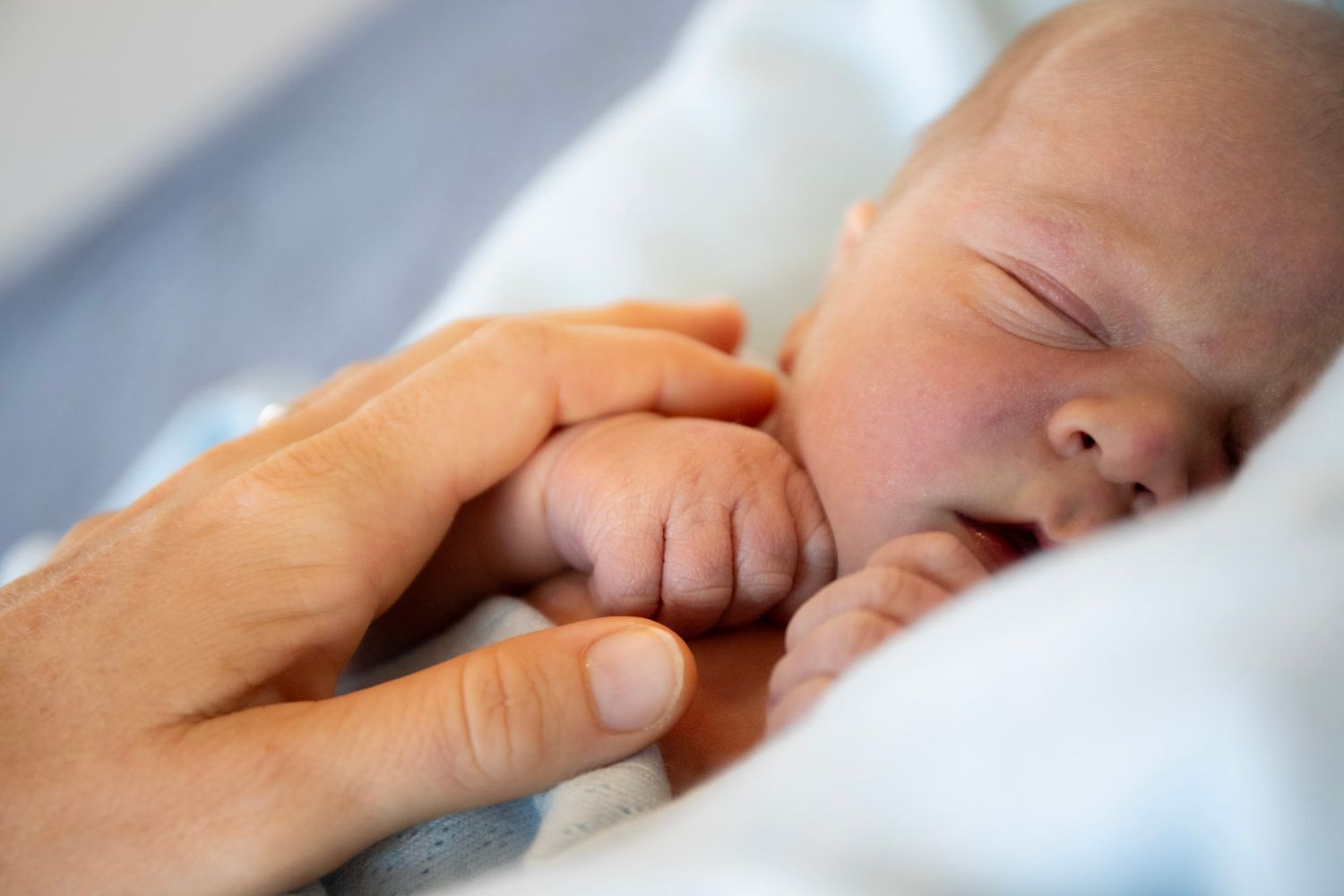 Newborn baby sleeping peacefully while gently gripping an adult's finger, wrapped in a soft white blanket.