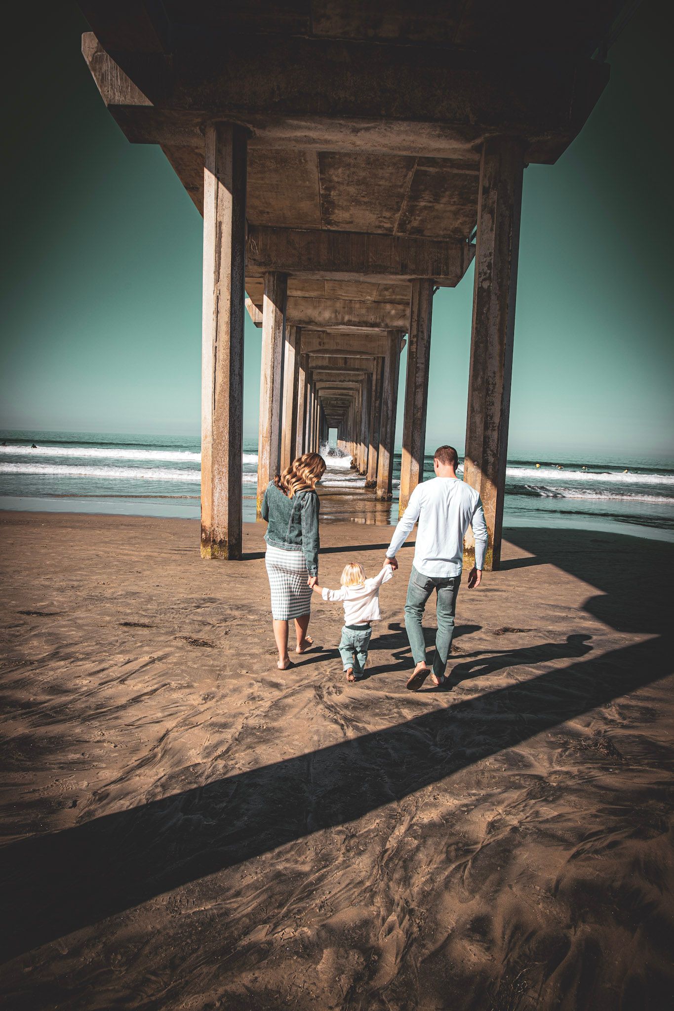 Family walking hand-in-hand under a beach pier on sandy shore with ocean waves in the background.