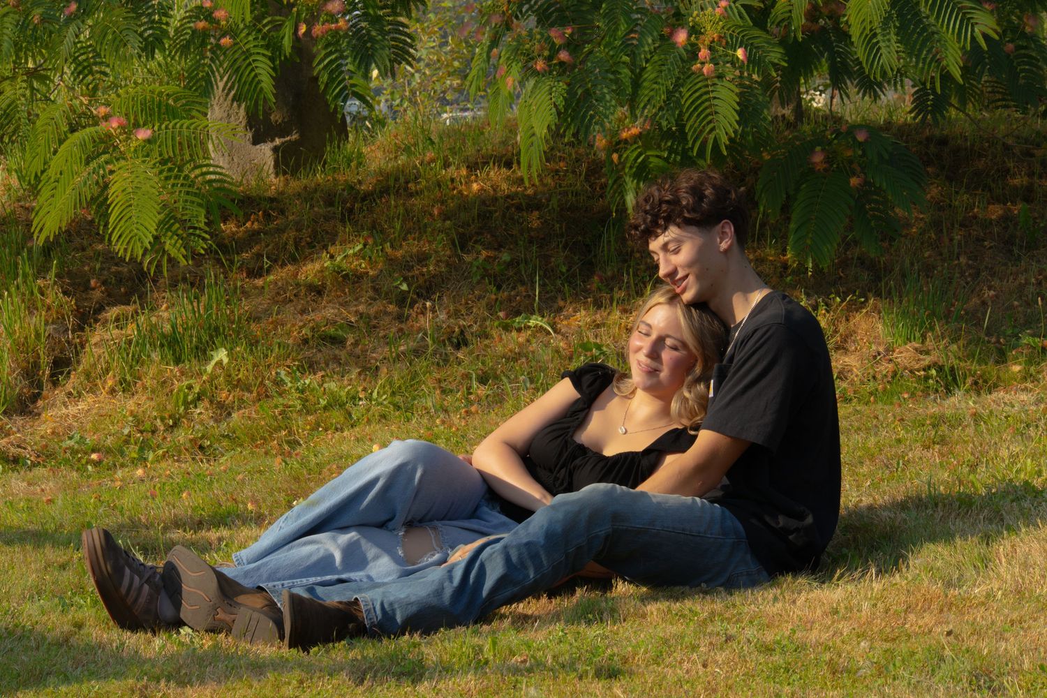 Young couple relaxing on green grass under a tree, enjoying a peaceful outdoor moment together on a sunny day.