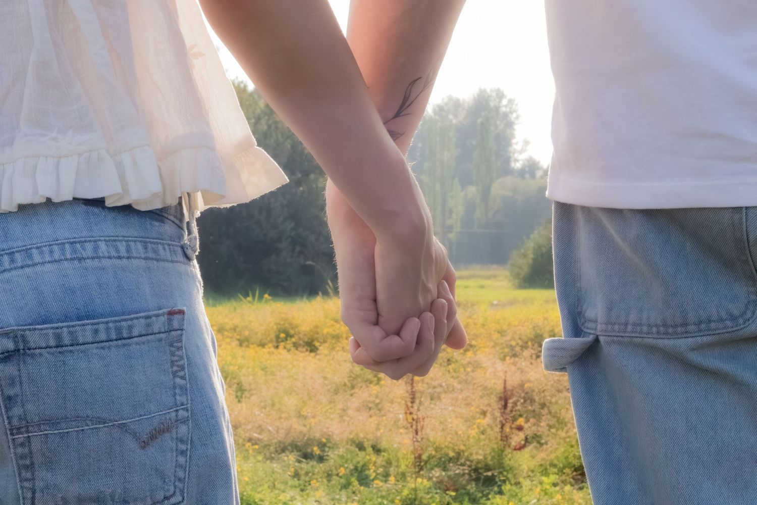 Couple holding hands outdoors in a sunny field, wearing jeans and white tops, romantic nature background.