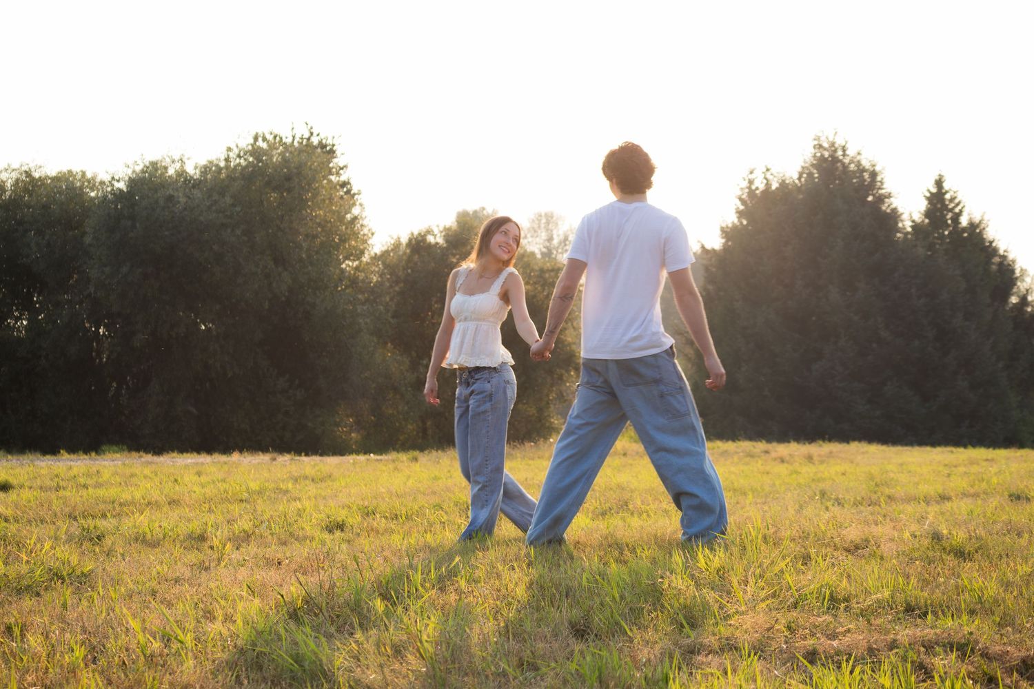 Couple holding hands walking in a sunny green meadow with trees in the background during golden hour.