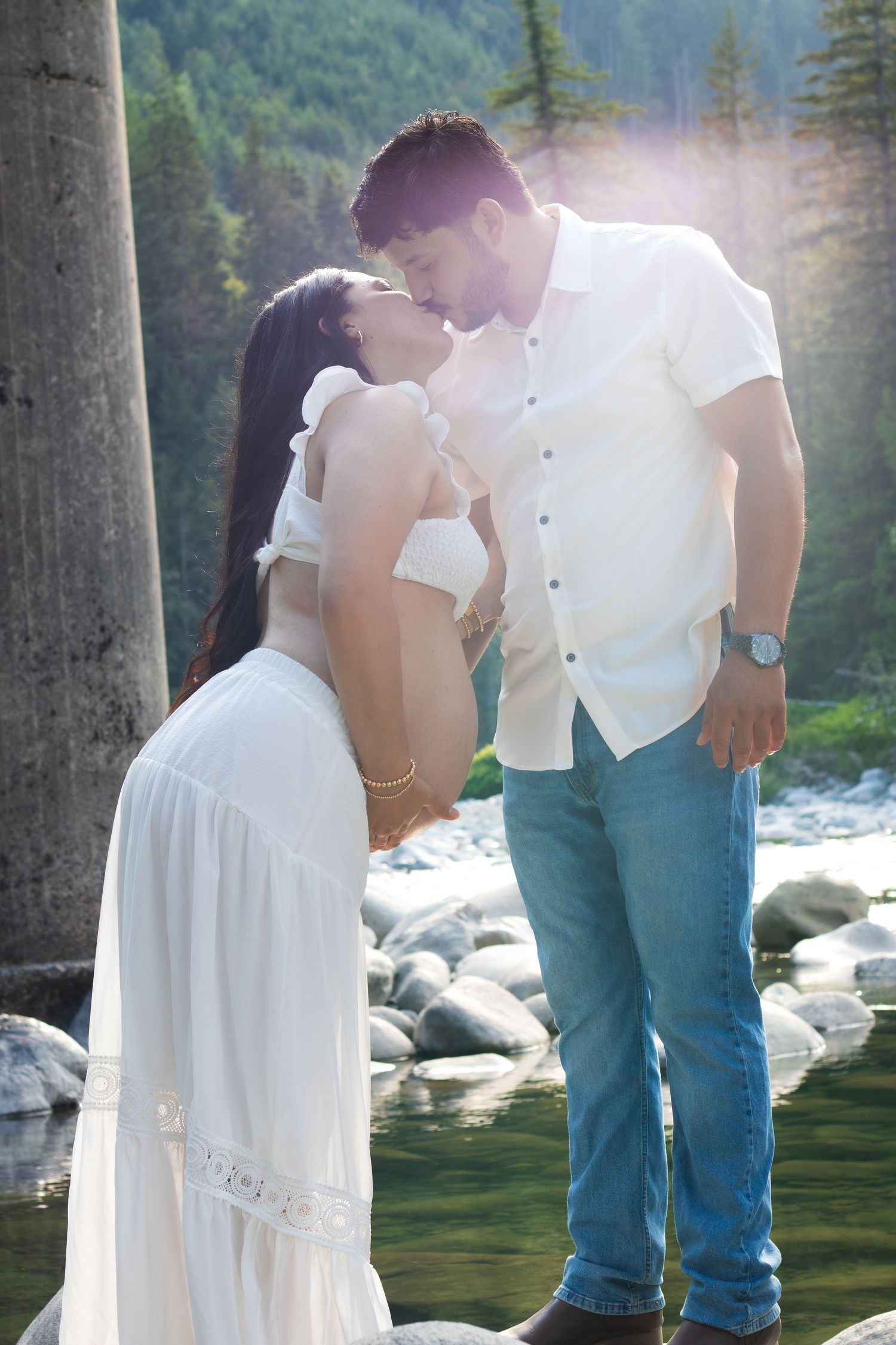 Couple sharing a romantic kiss by a rocky river, woman in white dress, man in white shirt and jeans, forest backdrop.