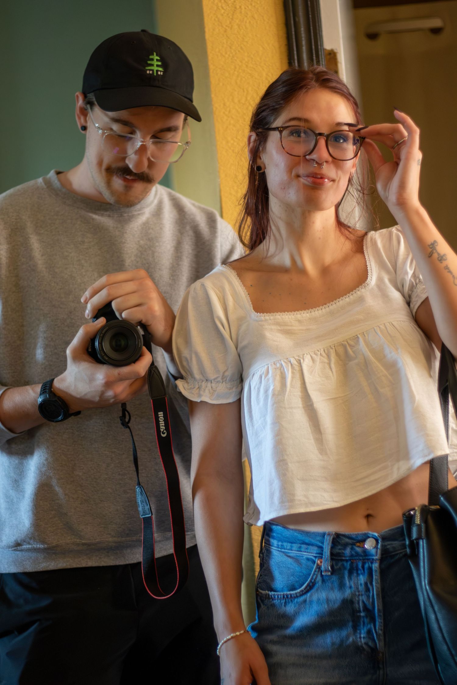 A young man with a Canon camera and a woman in a white crop top and glasses pose together indoors.