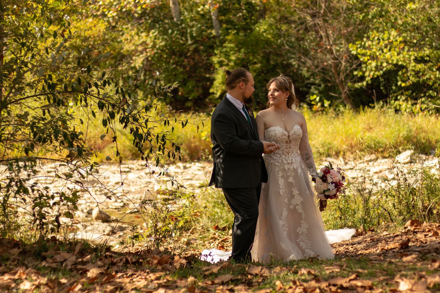 Bride and groom stand together near a sunlit creek surrounded by autumn foliage in a romantic outdoor wedding portrait.