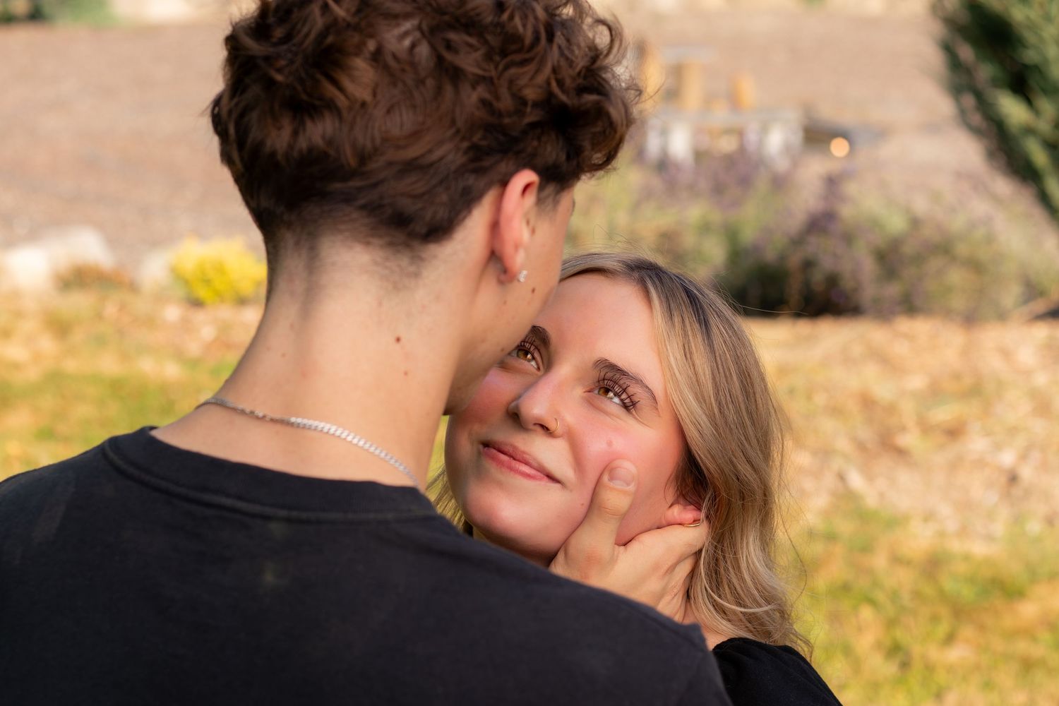 A young man tenderly holds a smiling blonde woman's face outdoors in warm sunlight.