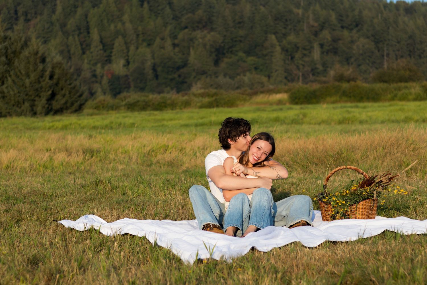 Couple embracing on white blanket during romantic outdoor picnic in green meadow with forest background.