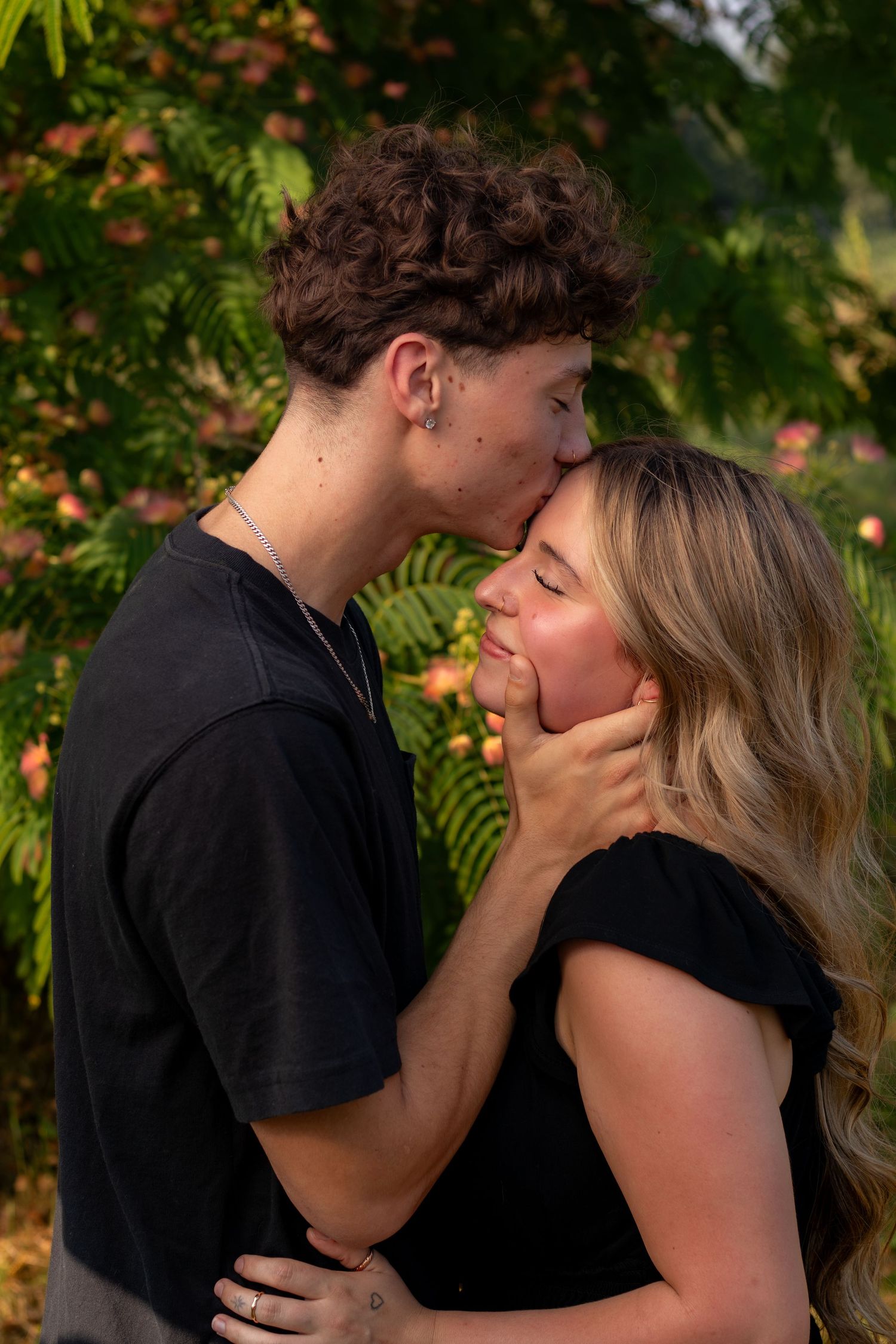 A young couple shares a tender forehead kiss outdoors, surrounded by lush green and pink foliage.