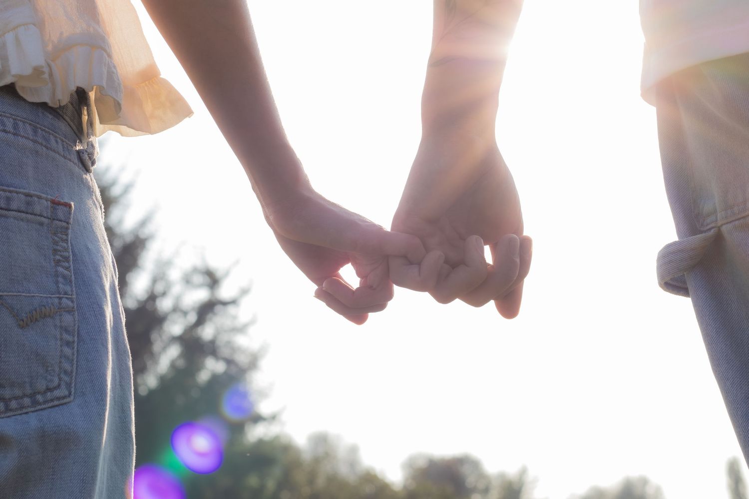 Two people holding hands with interlinked fingers outdoors in soft sunlight, wearing casual jeans.