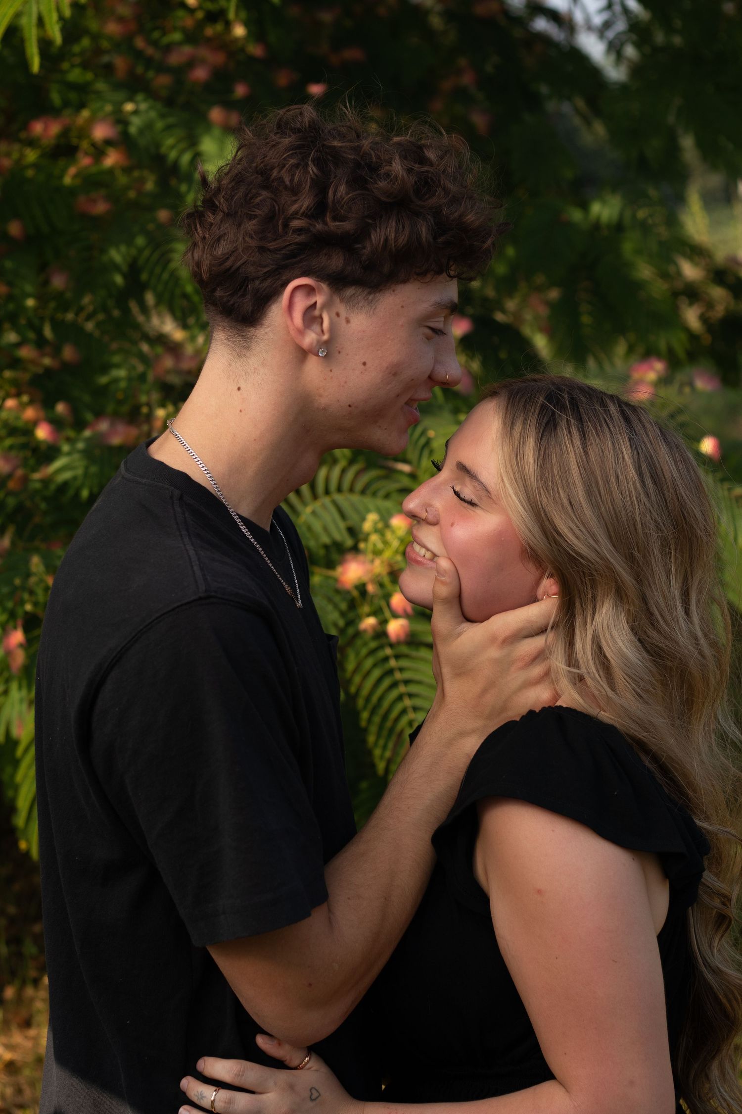 A young couple in black tops share a romantic moment outdoors, with the man gently holding the womans face near a flowering tree.