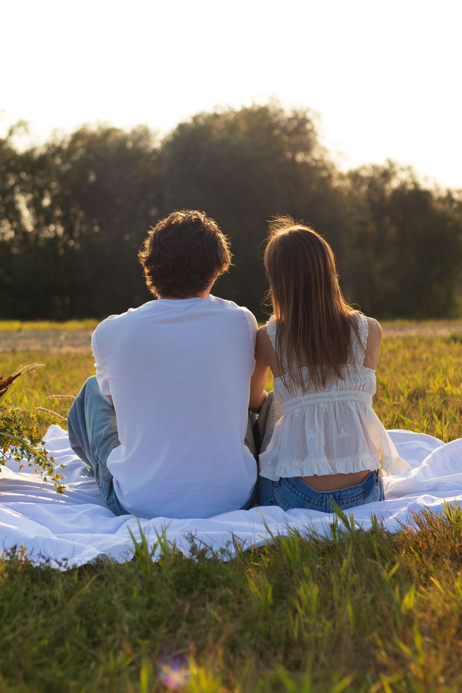 Couple sitting on a white blanket in a sunny field at sunset, viewed from behind, surrounded by green grass.