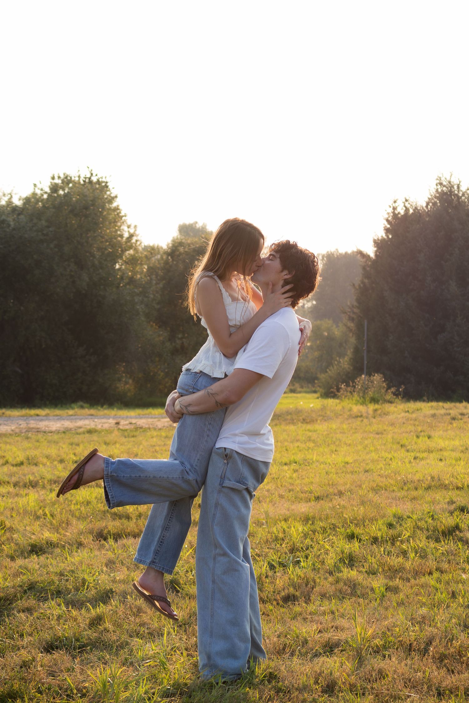 Couple sharing a romantic kiss in a sunlit green field, man lifting woman, surrounded by trees at golden hour.
