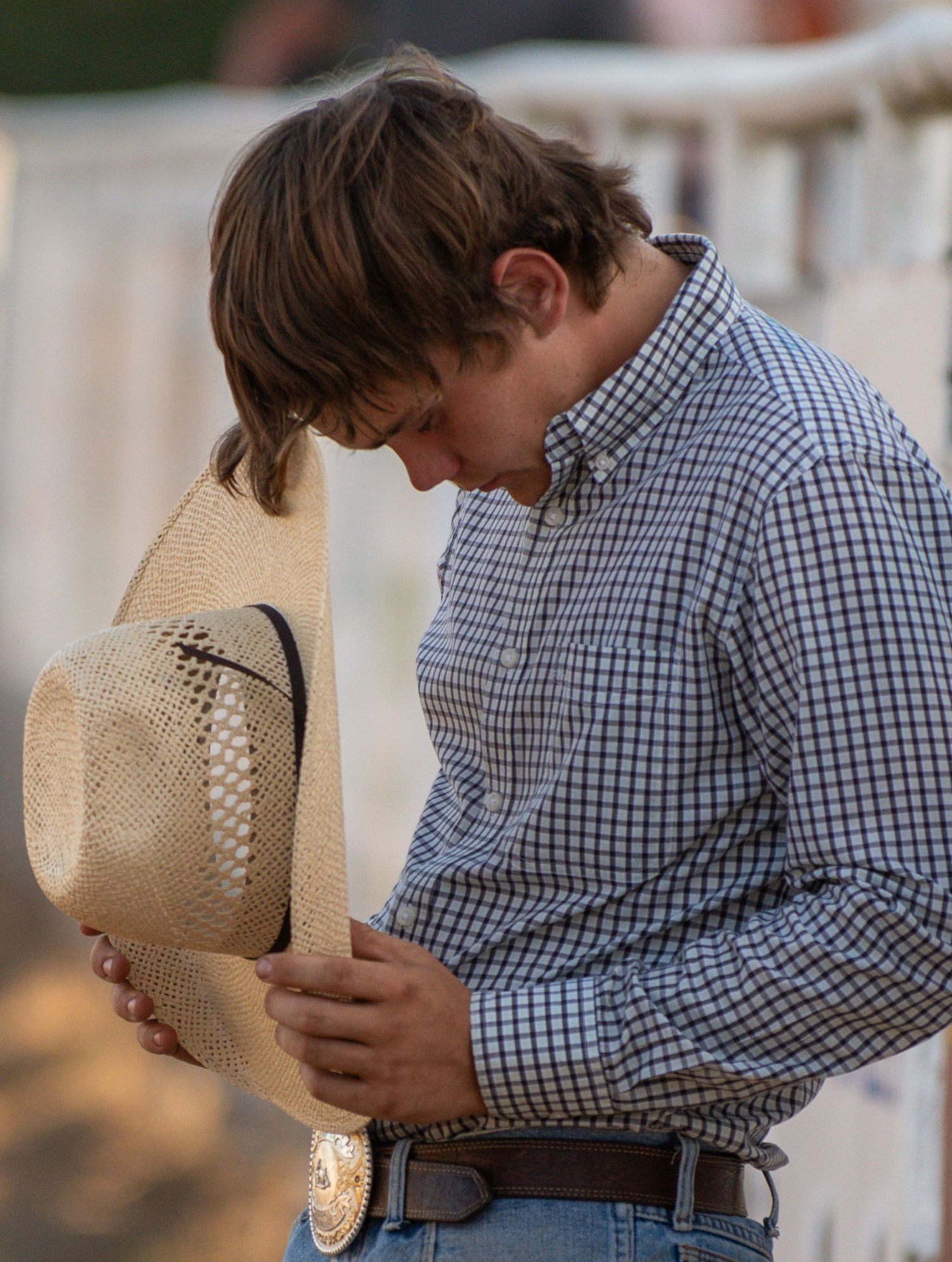 Young cowboy in plaid shirt bowing his head while holding a straw cowboy hat with a belt buckle.