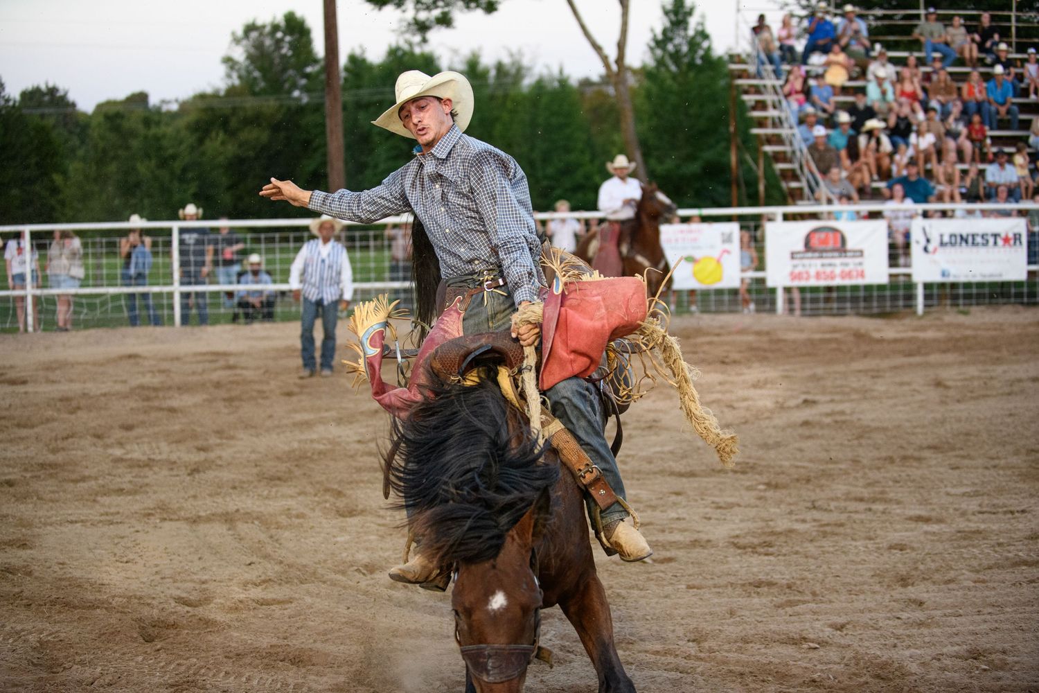 Cowboy riding a bucking bronco at a rodeo competition with spectators watching in the background.
