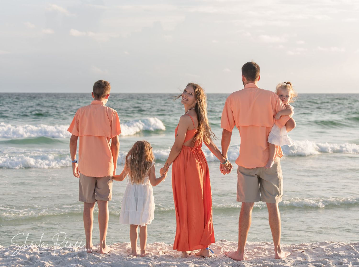 Family of five holding hands on a white sand beach at sunset, wearing coordinating coral and white outfits.