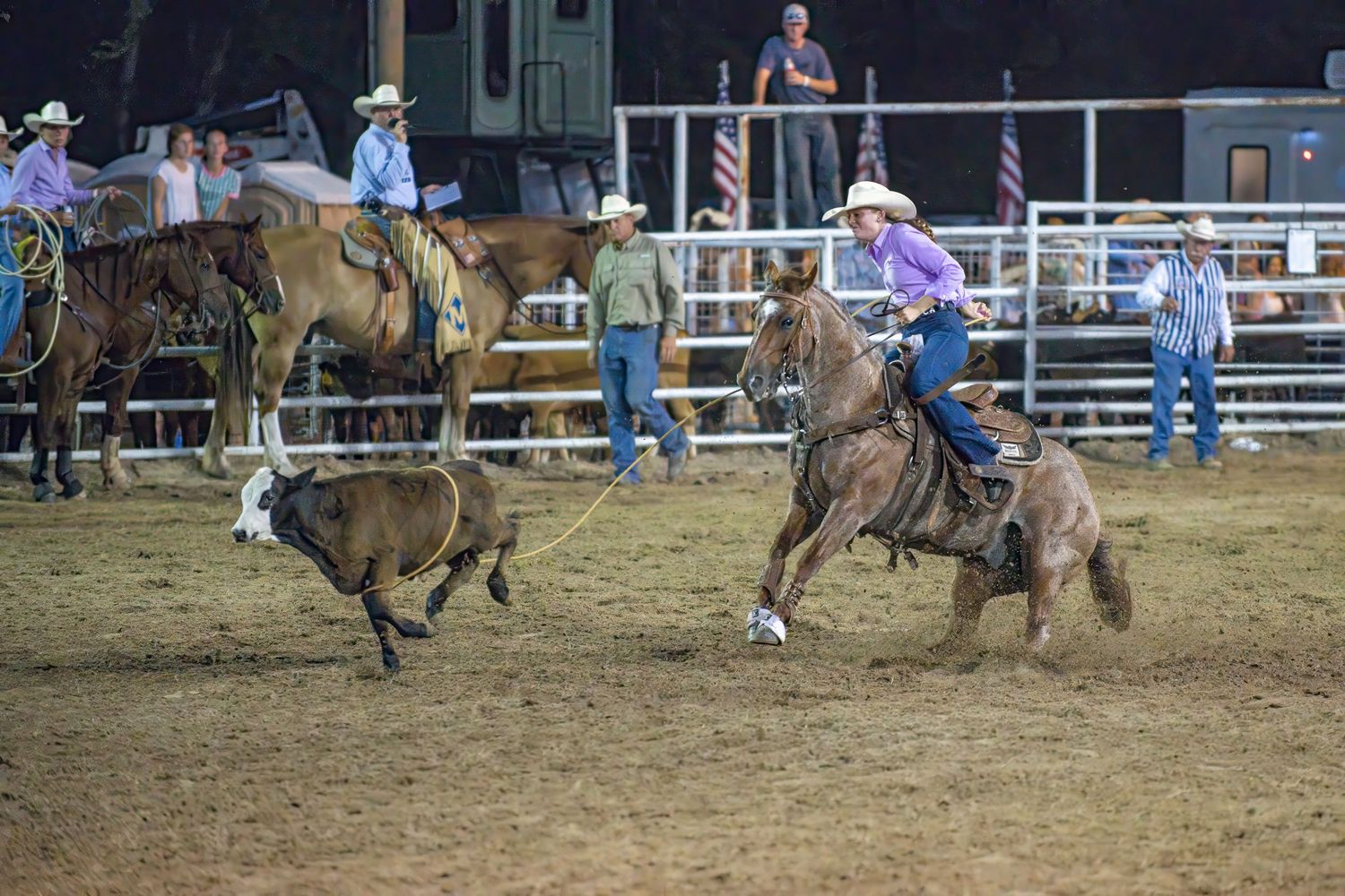 Cowboy on horseback chasing a calf with a lasso during a nighttime rodeo roping competition.