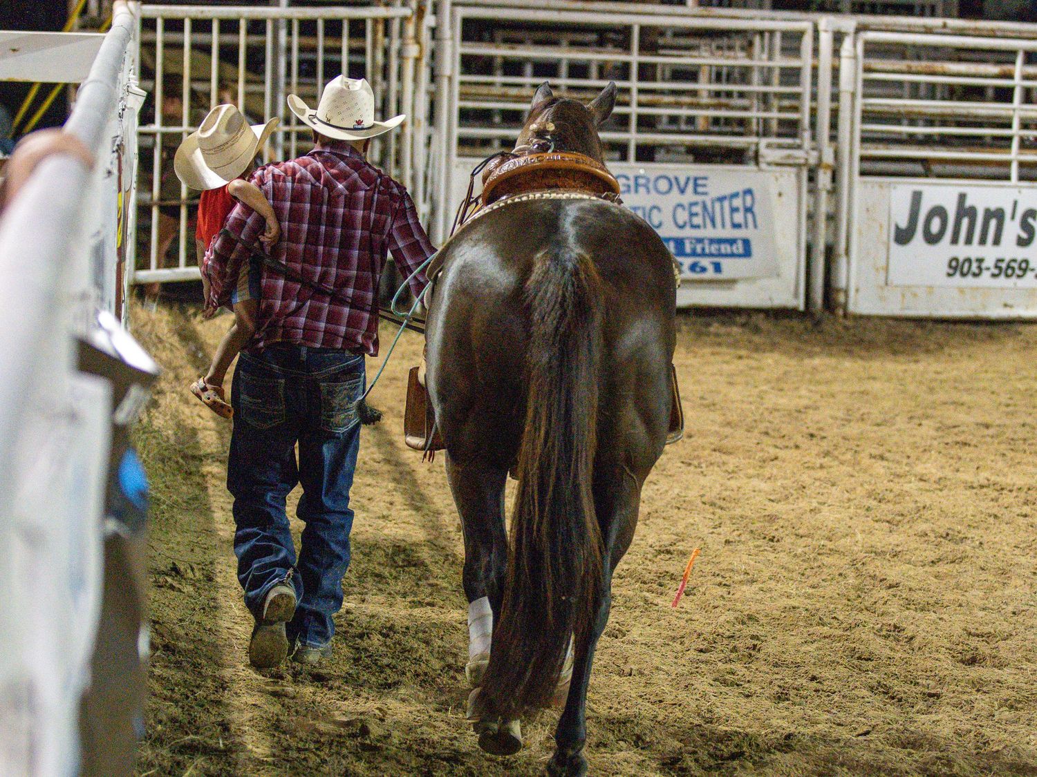 Two cowboys walk a brown horse through an indoor rodeo arena dirt floor.