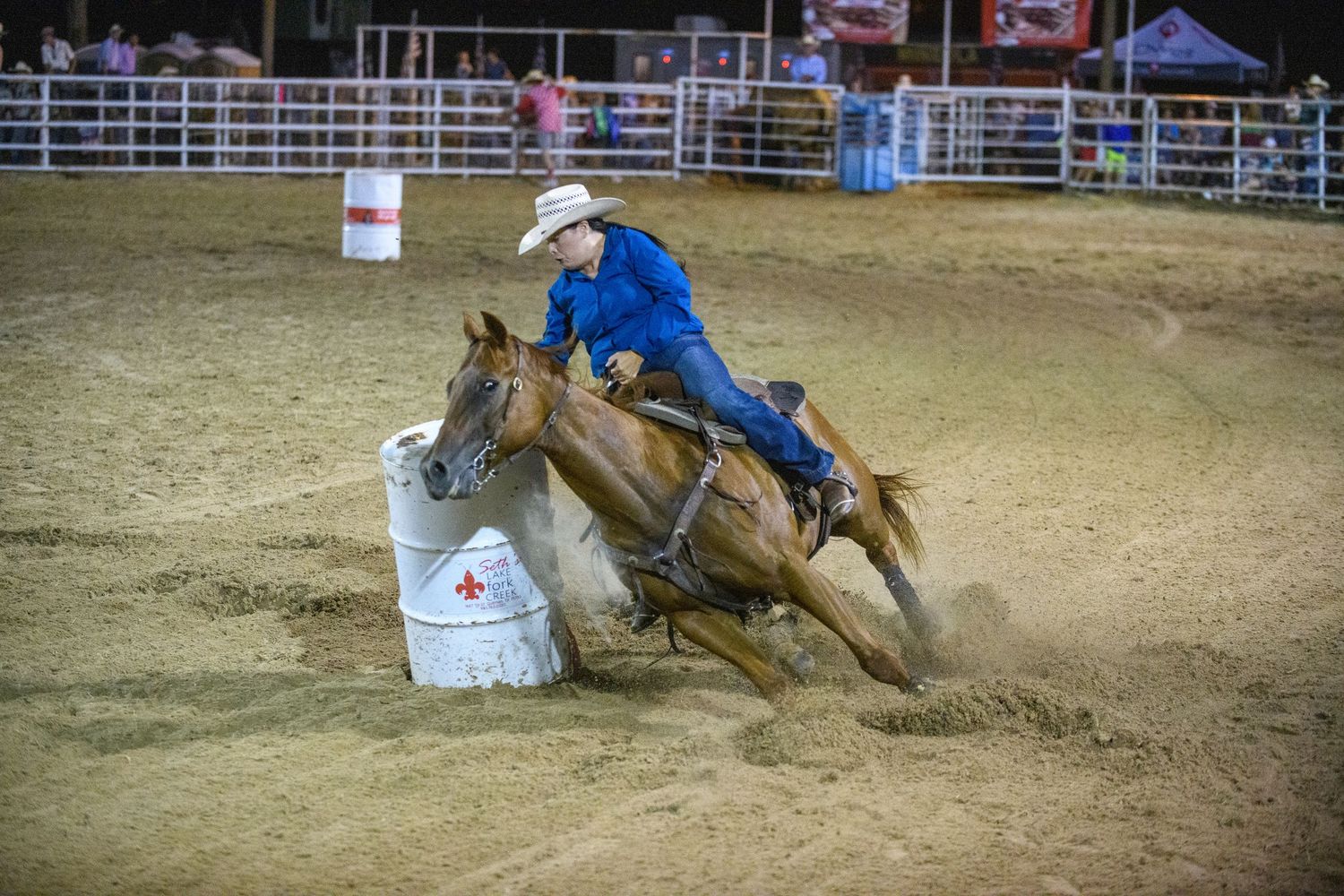 Rodeo competitor in blue shirt riding a horse around a barrel during an indoor barrel racing event.