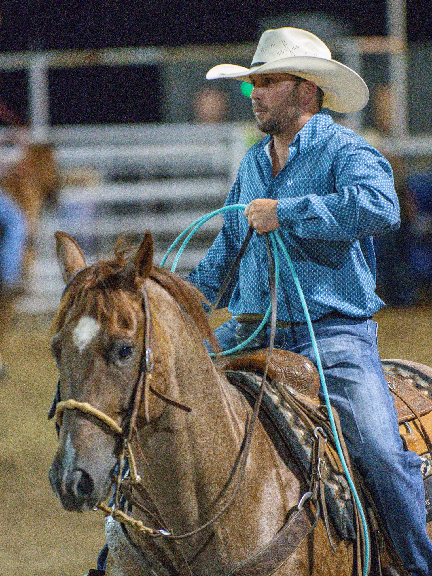 Cowboy in blue shirt on horseback holding a blue lasso rope at a rodeo event.